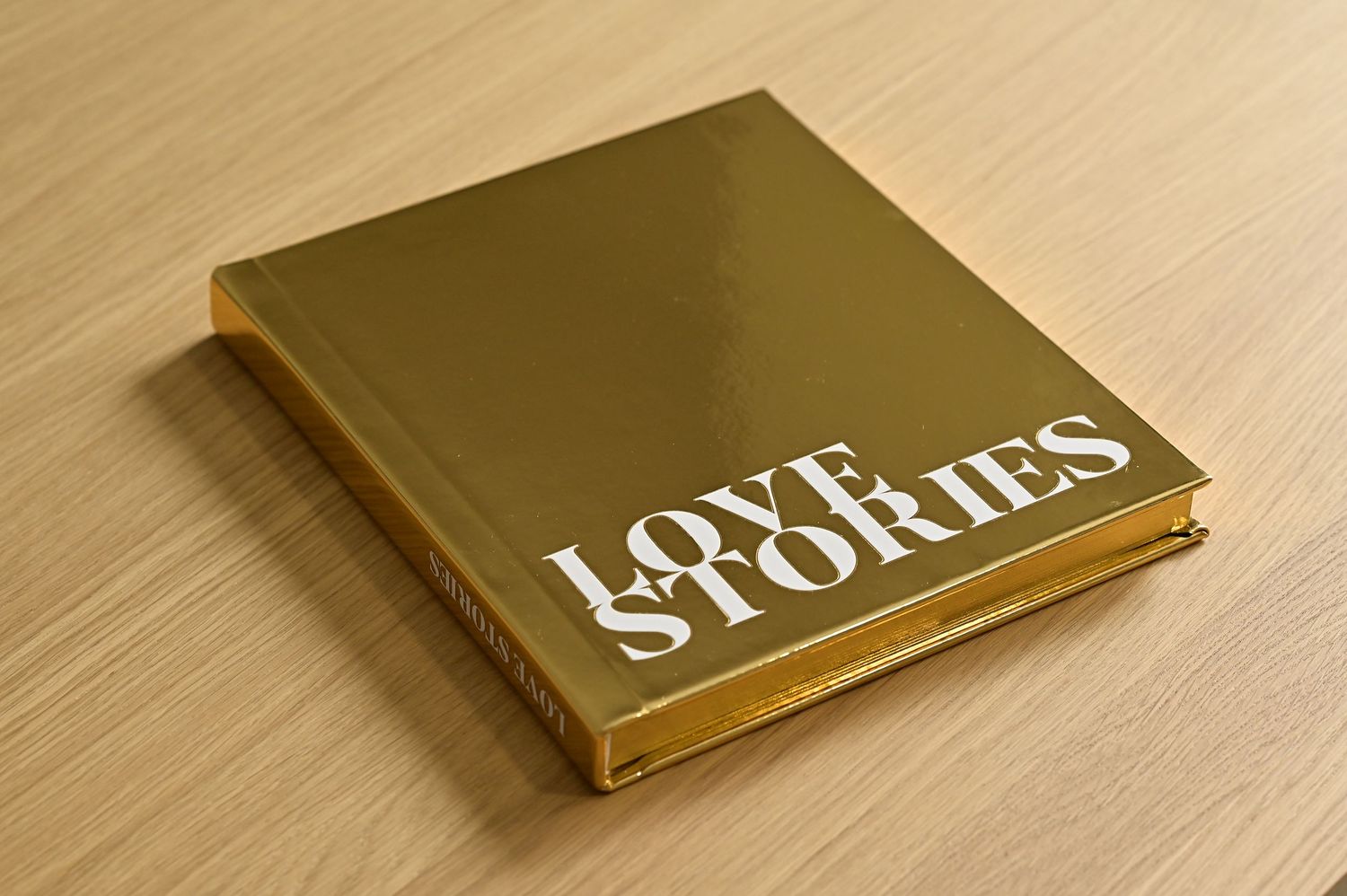 Gold metallic Love Stories wedding photo album with white typography on a light wooden surface.