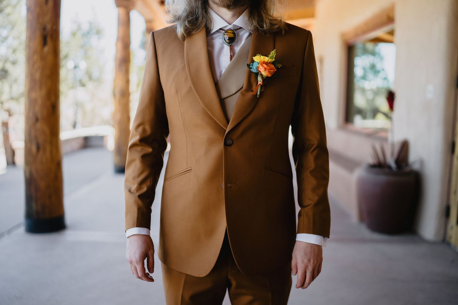 Groom in a caramel brown suit with colorful boutonniere at an outdoor wedding venue.