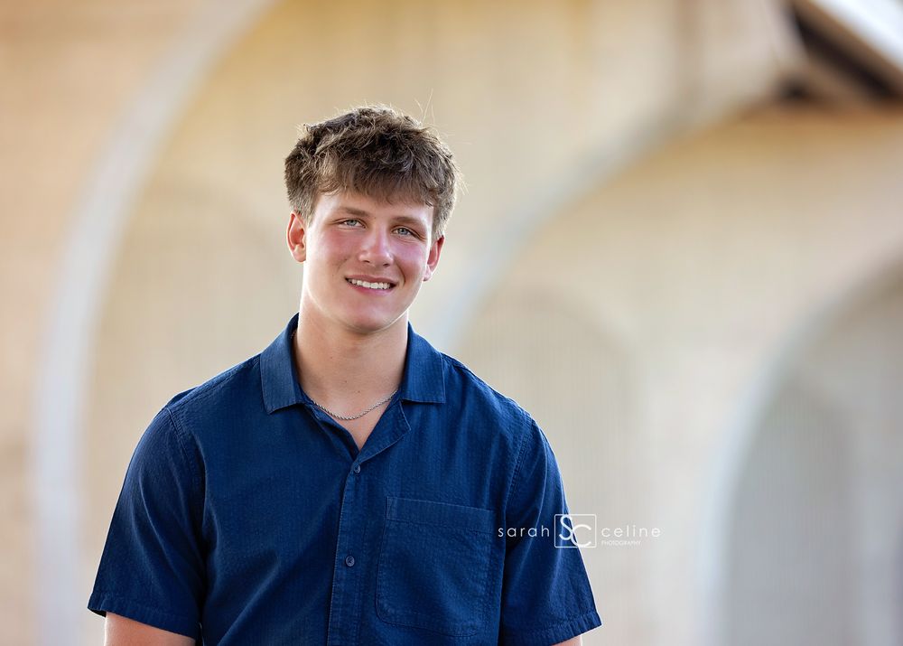 Smiling teenage boy in navy blue shirt poses outdoors under arched architecture for senior portrait photo.