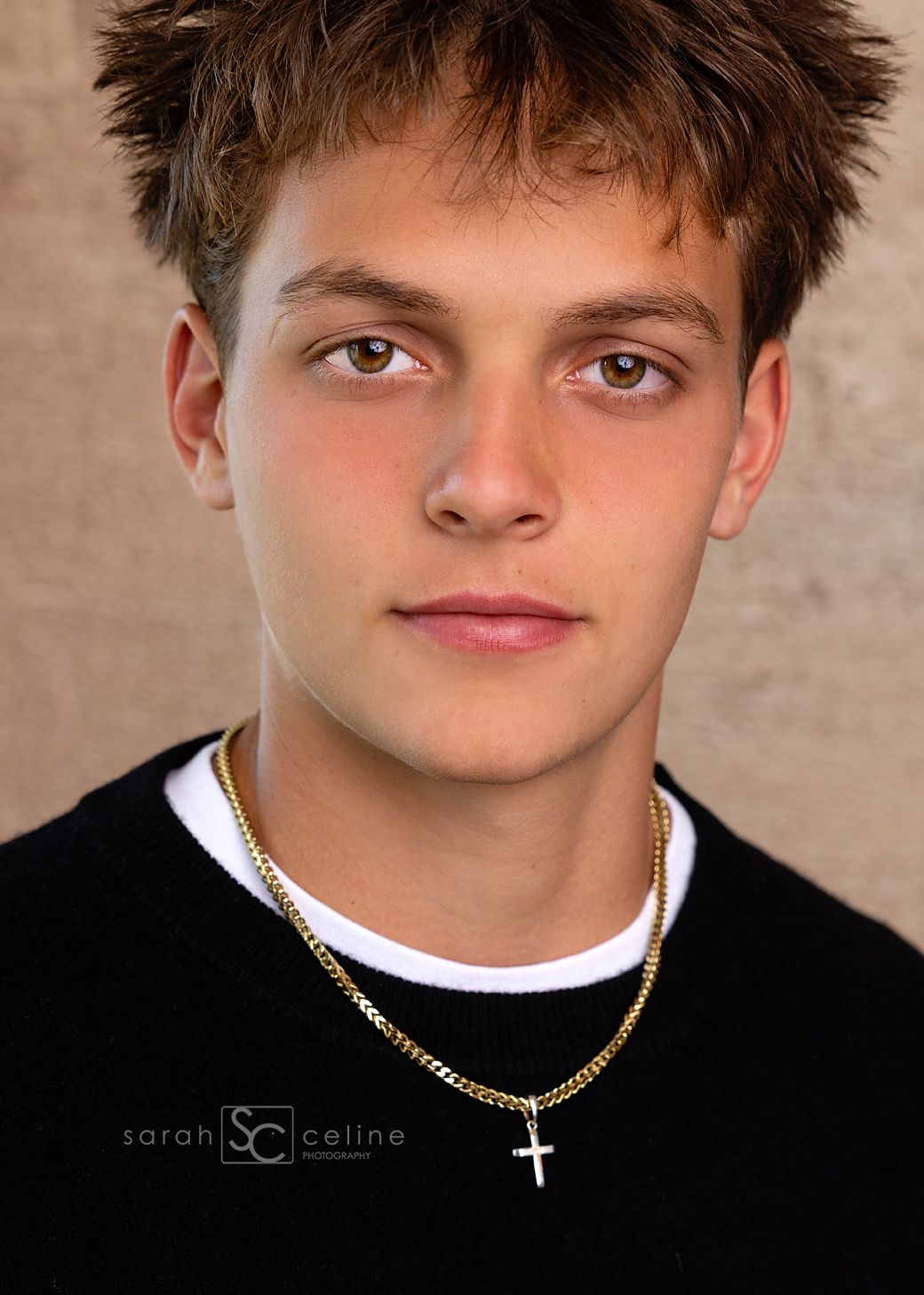 Young man with brown hair wearing a black sweater and gold cross necklace poses for a professional portrait photo.