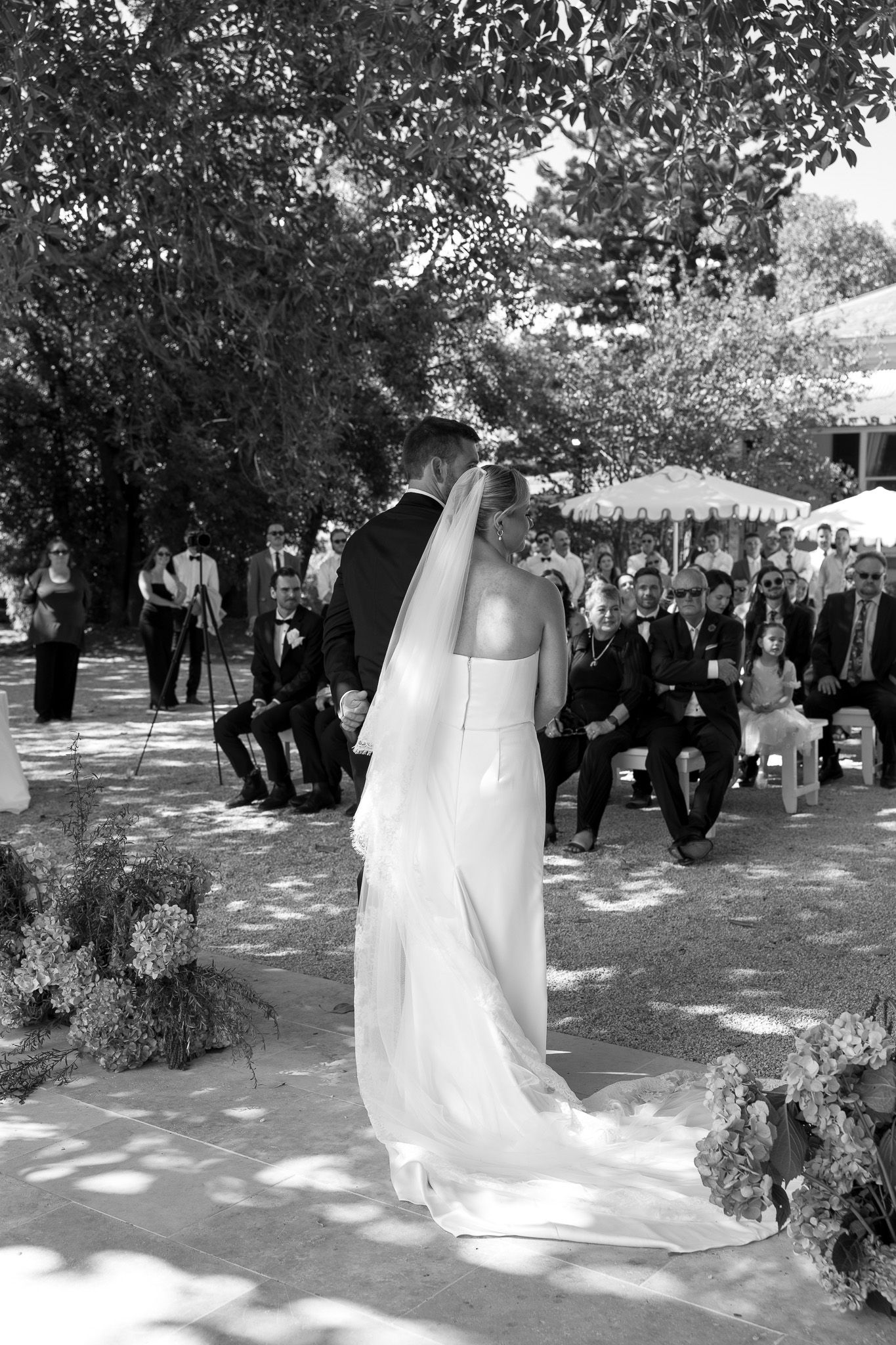 Black and white photo of a bride and groom at an outdoor wedding ceremony with seated guests under shady trees.