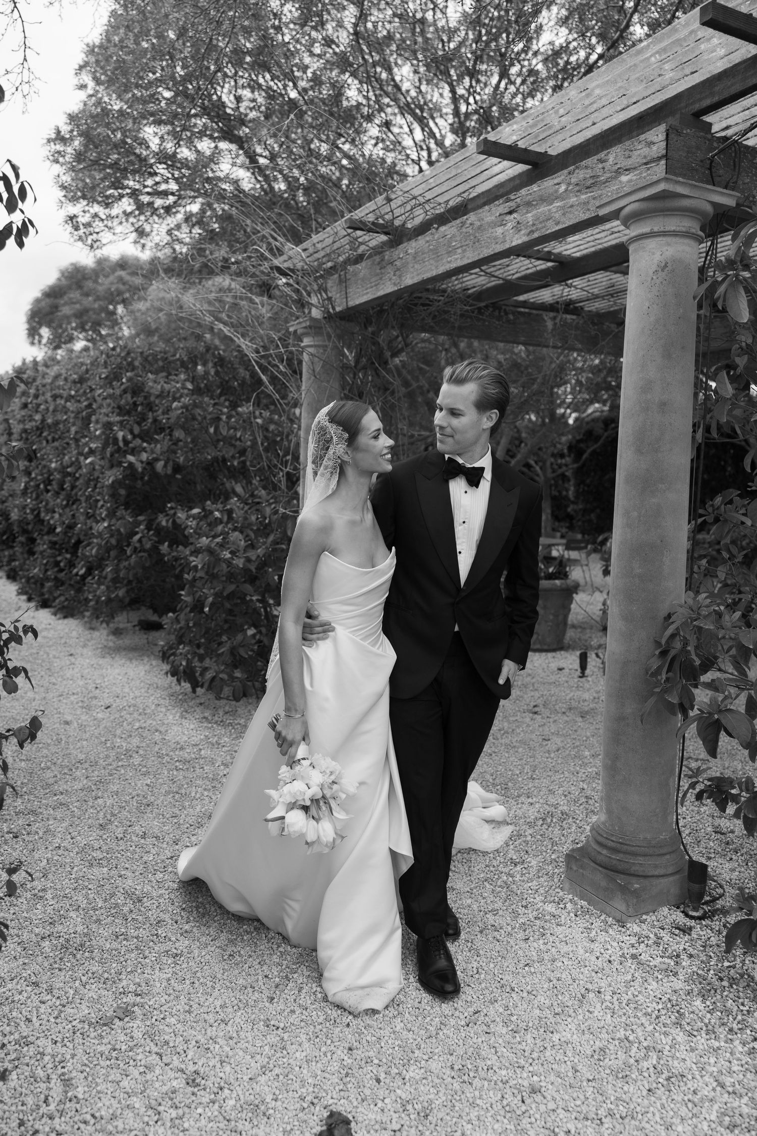 Black and white photo of a bride and groom at an outdoor wedding ceremony with seated guests under shady trees.