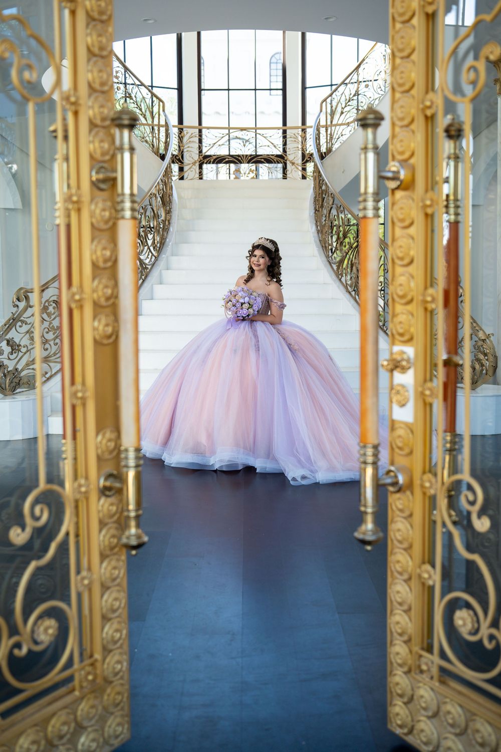 Young woman in a lavender ball gown holds a bouquet at the base of grand white stairs with ornate gold railings.