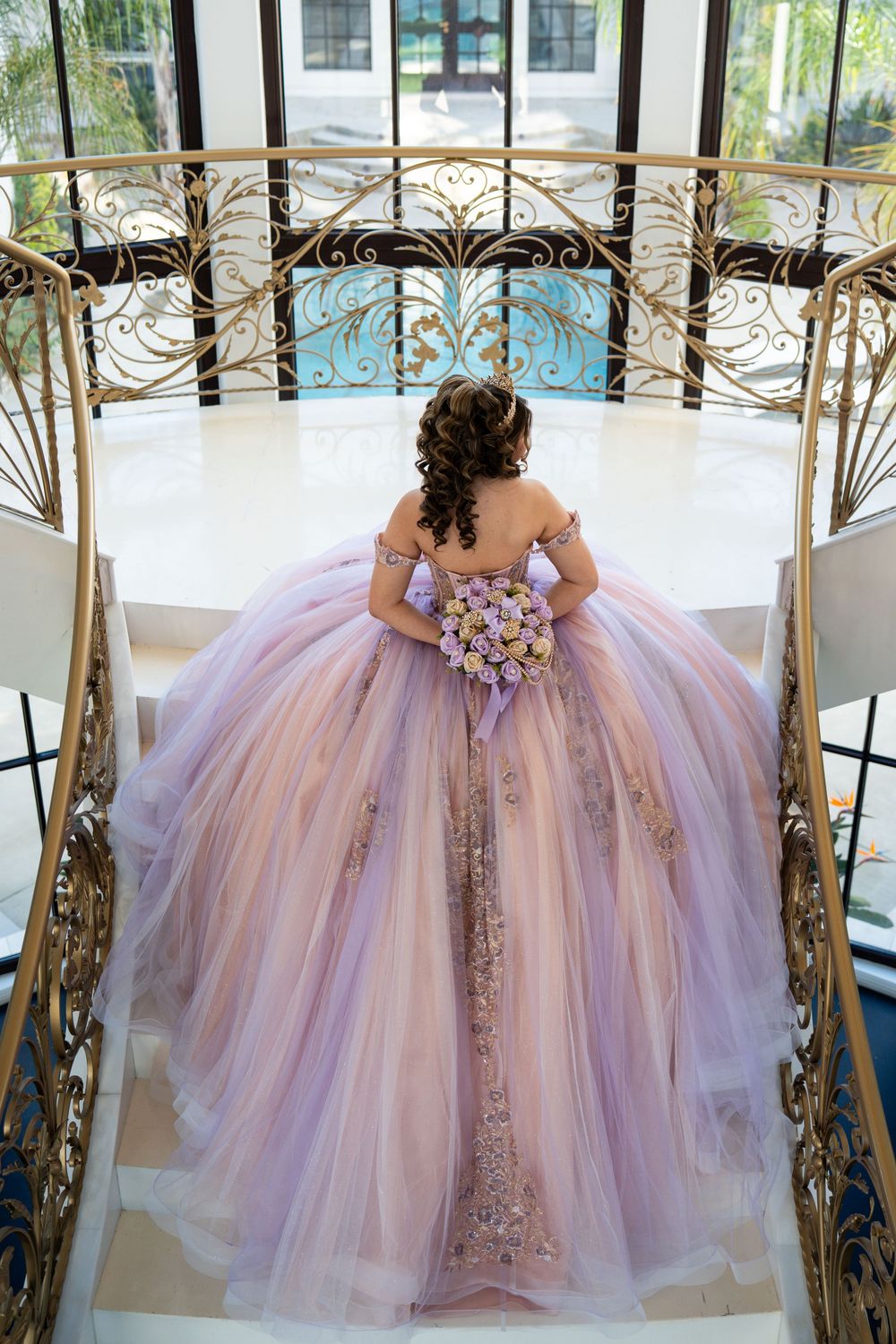 Back view of a woman in a lavender quinceañera ball gown with gold embroidery on elegant white marble stairs.