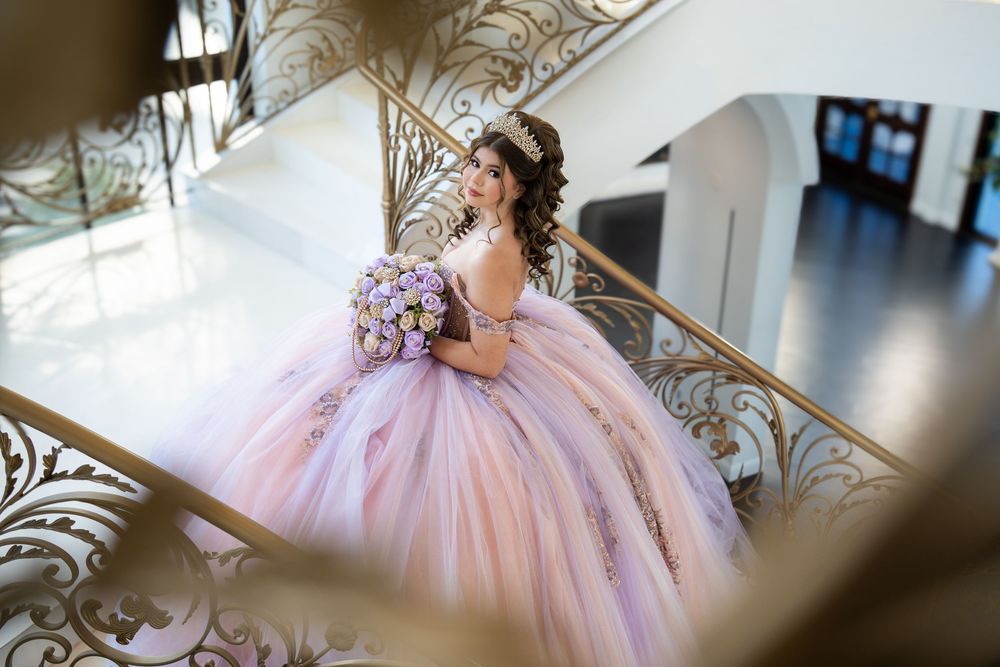 Young woman in a pink ball gown holding a bouquet on an elegant staircase during her quinceañera celebration.