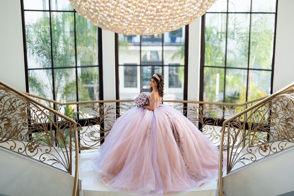 Young woman in a pink ball gown quinceañera dress poses on elegant staircase with ornate railings and chandelier.