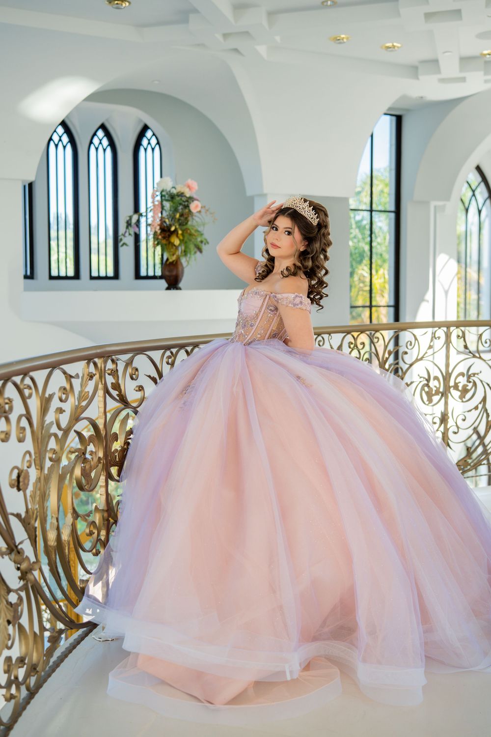 Young woman in pink ball gown with tiara poses on ornate golden staircase in elegant white venue.