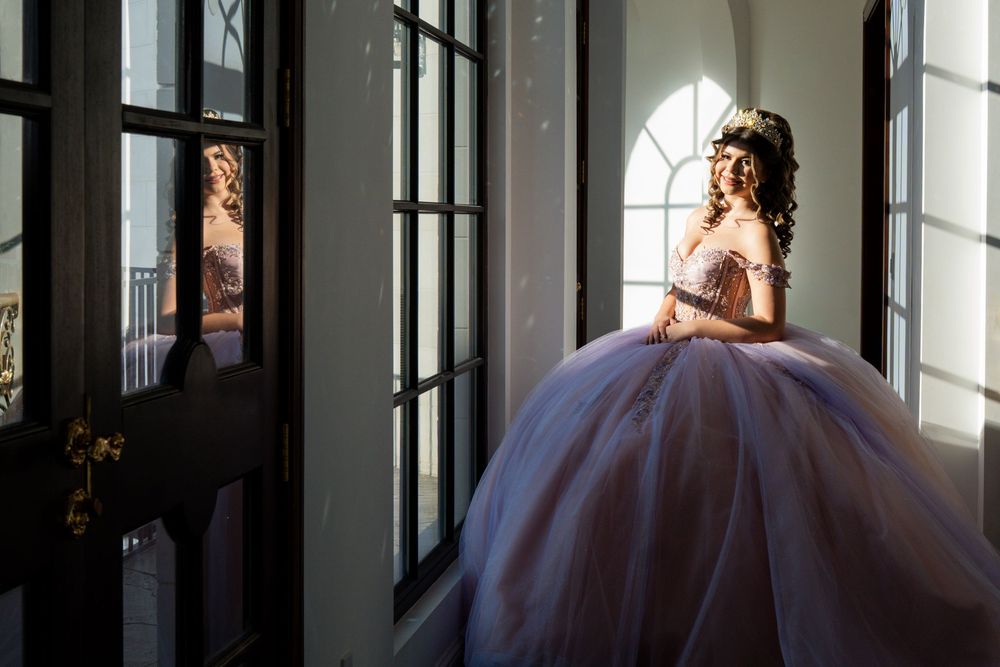Young woman in a lavender ball gown quinceañera dress stands by elegant French doors in soft natural light.