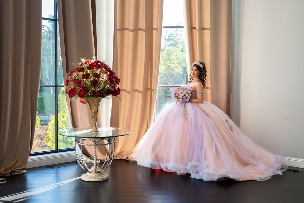 Young girl in a pink ball gown quinceañera dress poses by large windows with red roses in a vase nearby.