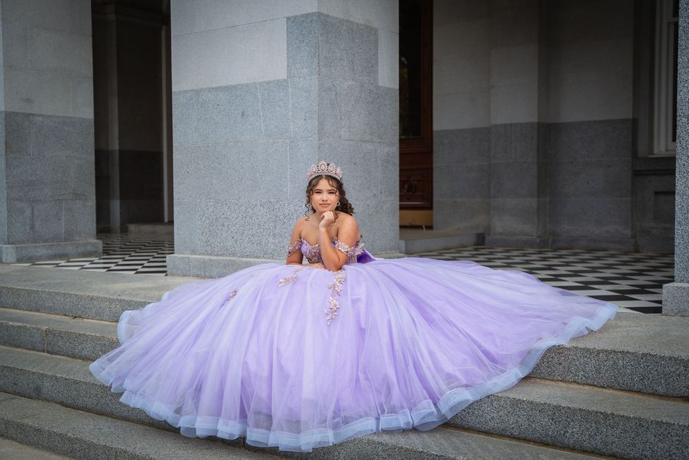 Teen girl in lavender quinceañera gown with crystal crown seated gracefully on stone steps outdoors.