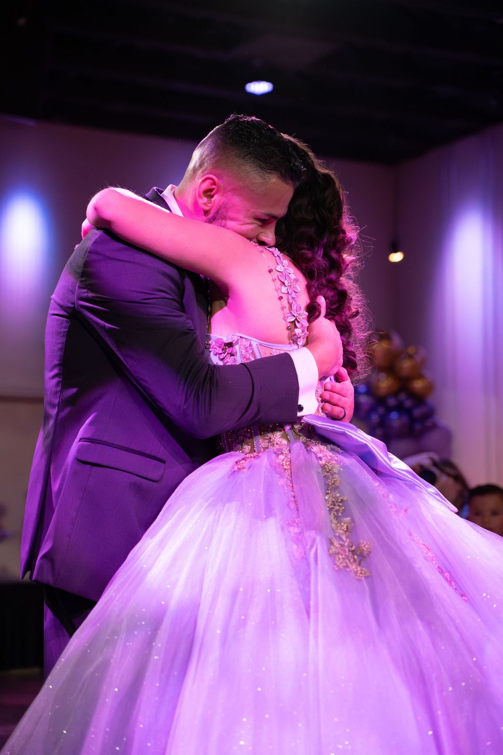 A quinceañera girl in a lavender ball gown shares an emotional dance embrace with a man in a purple suit.