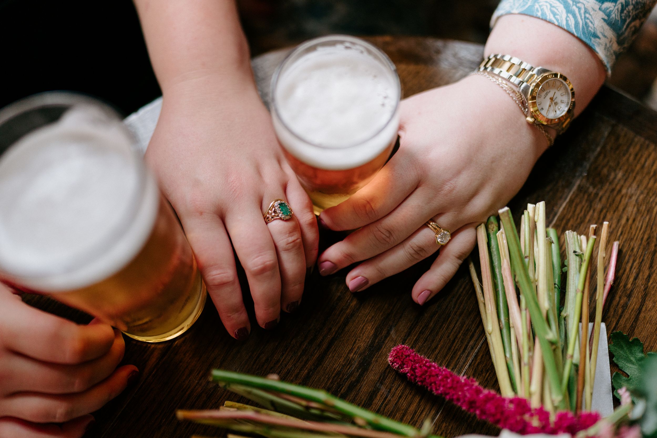 Hands holding frothy beer mugs on a wooden table with flowers and straws nearby, one wearing a gold Rolex watch.
