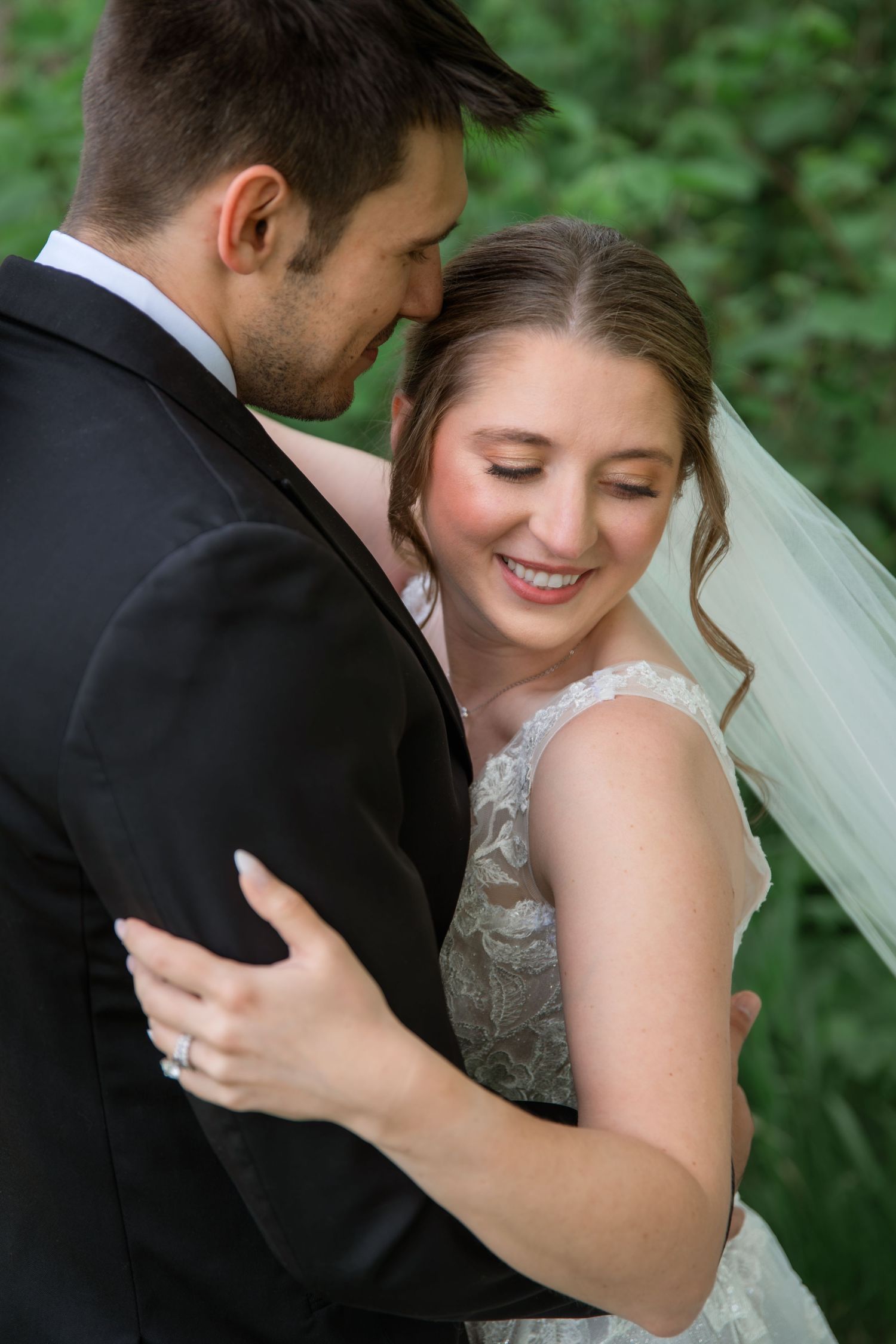 Bride in lace gown and veil smiles joyfully while embracing groom in black suit outdoors among green foliage.