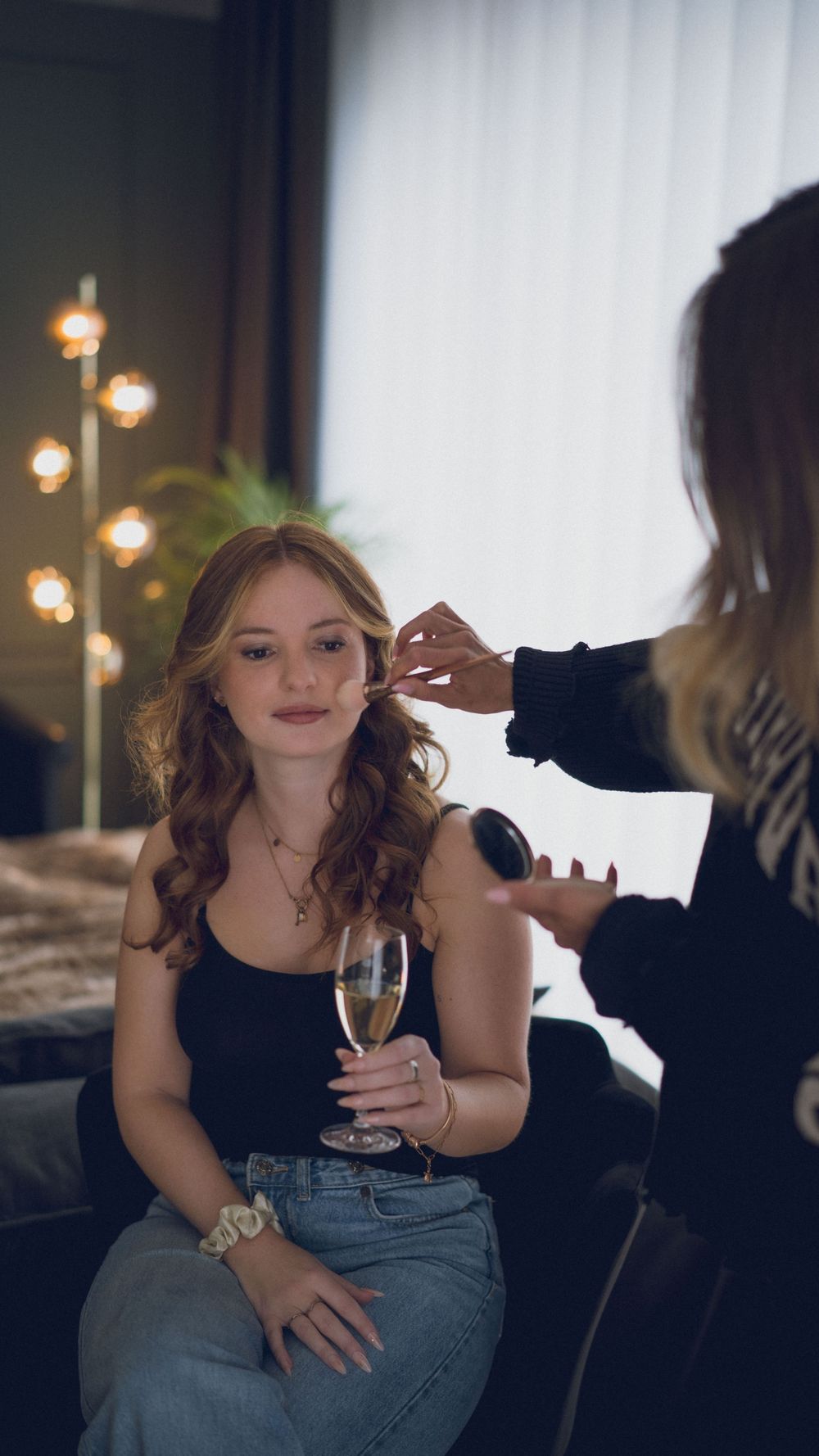 Junge Frau erhält professionelles Hair & Make-up und hält ein Glas Sekt vor ihrem Boudoir-Fotoshooting.