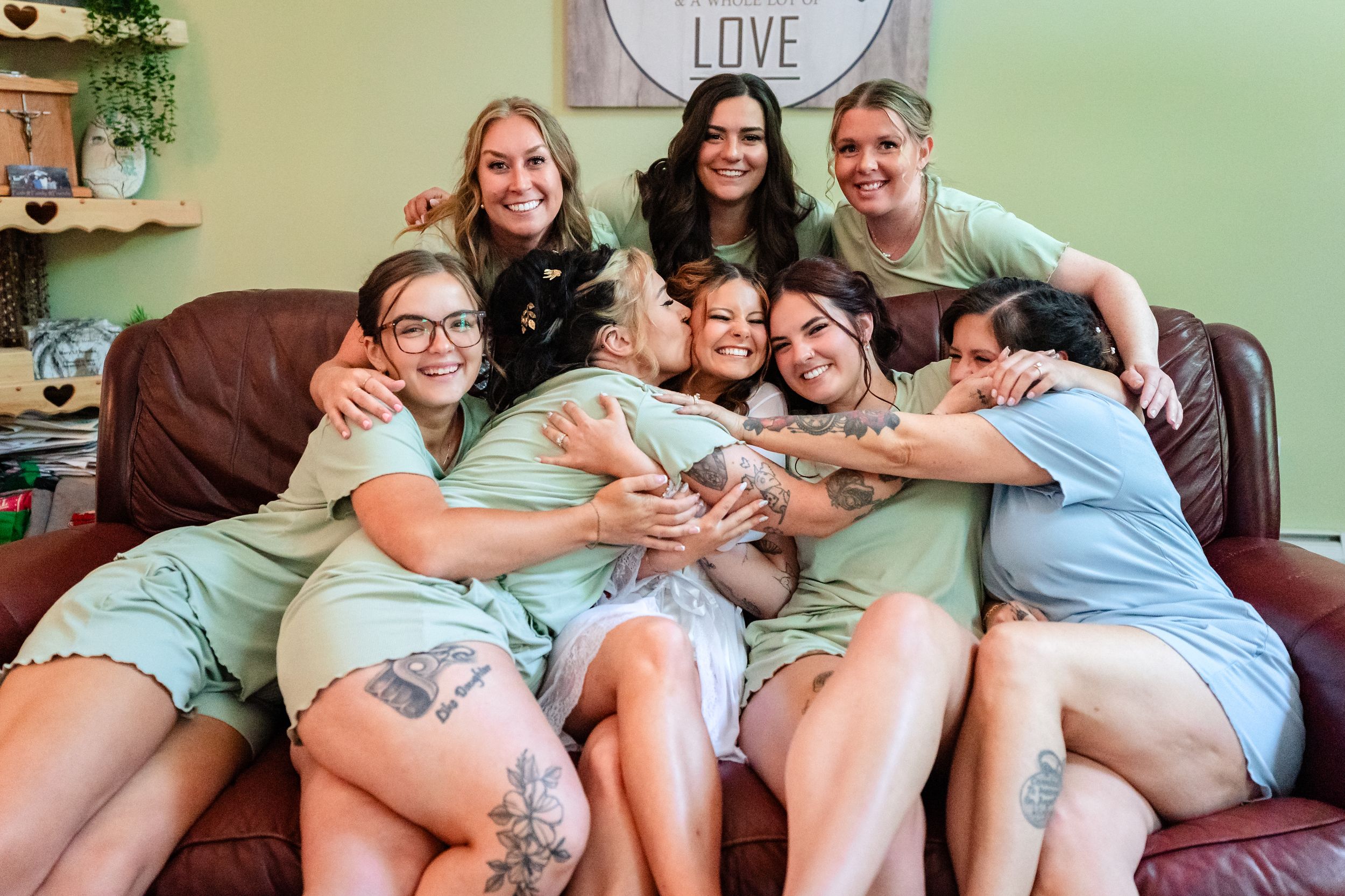 Bride surrounded by bridesmaids in sage green robes hugging on brown leather couch at getting ready.