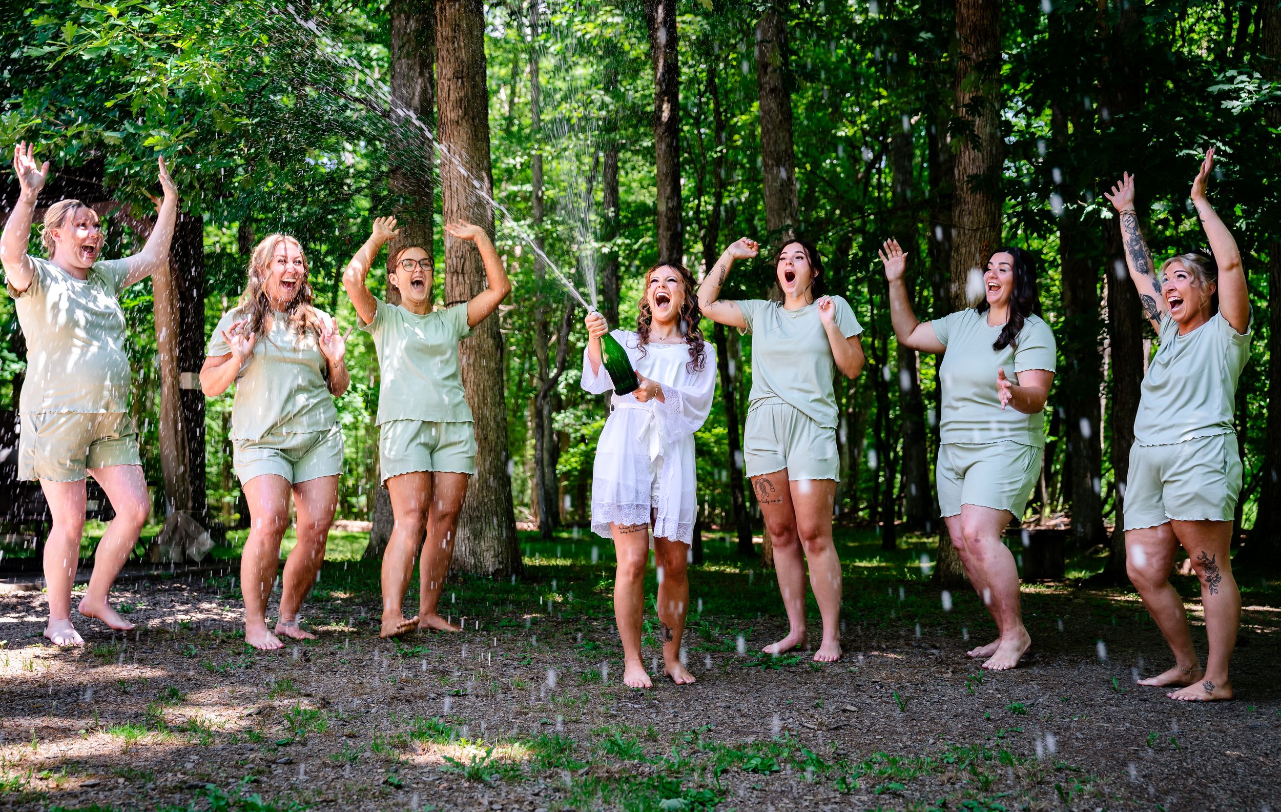 Excited bridal party in matching green rompers celebrating outdoors in lush forest setting.