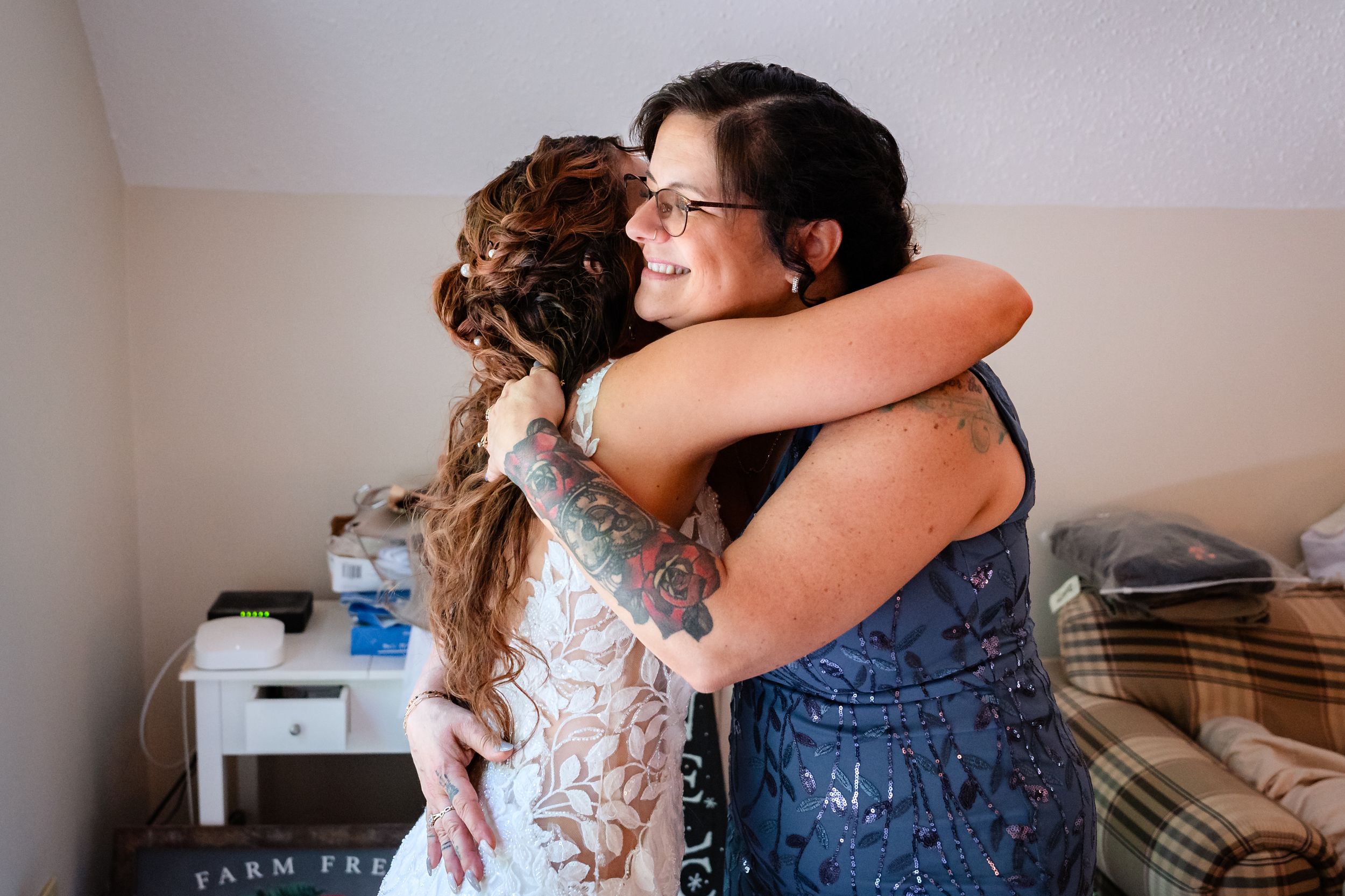 Emotional bride in wedding dress sharing warm hug with tattooed woman during wedding preparations.