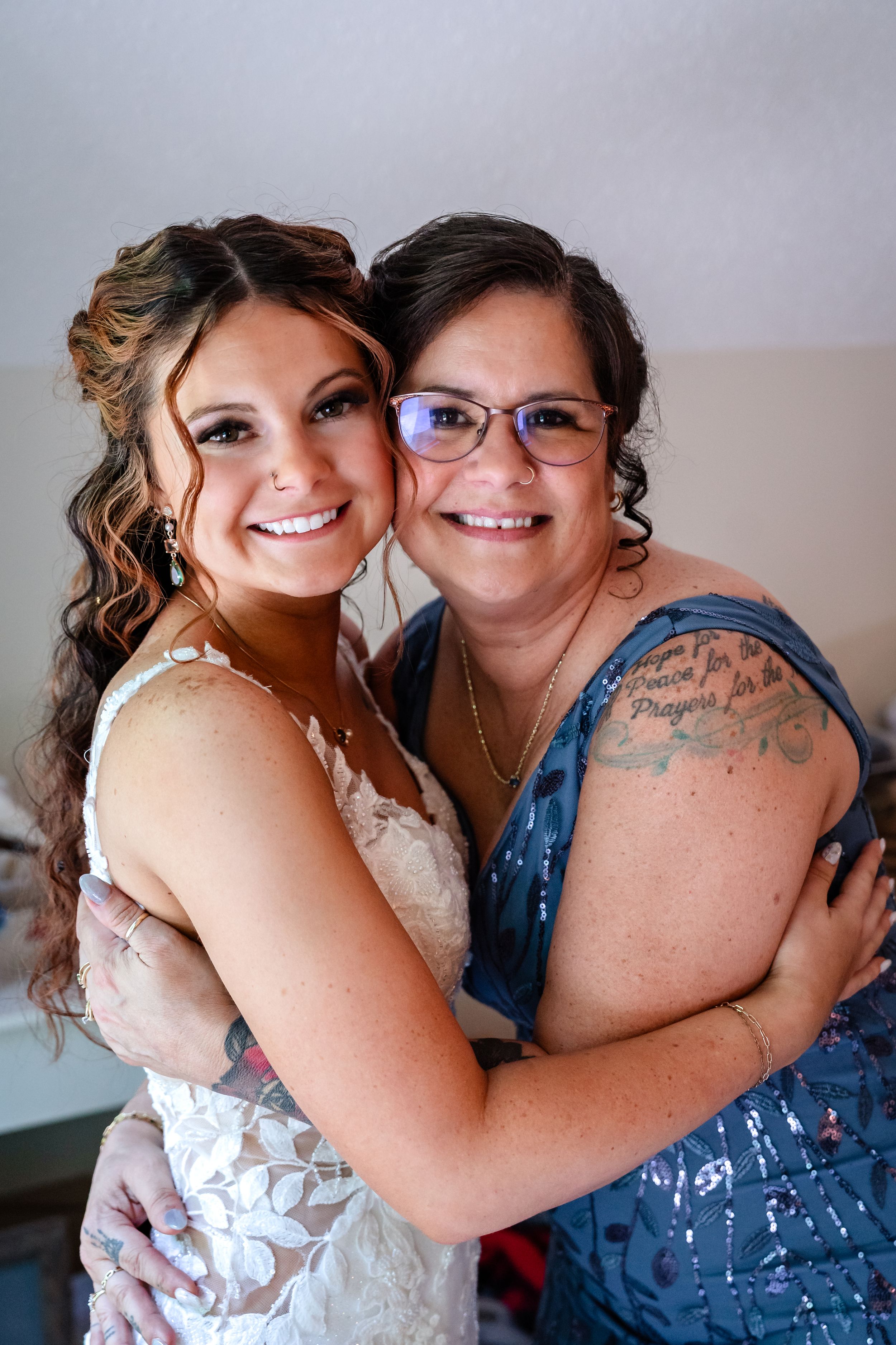 Bride and older woman in blue sequin dress share a warm embrace while smiling at the camera.