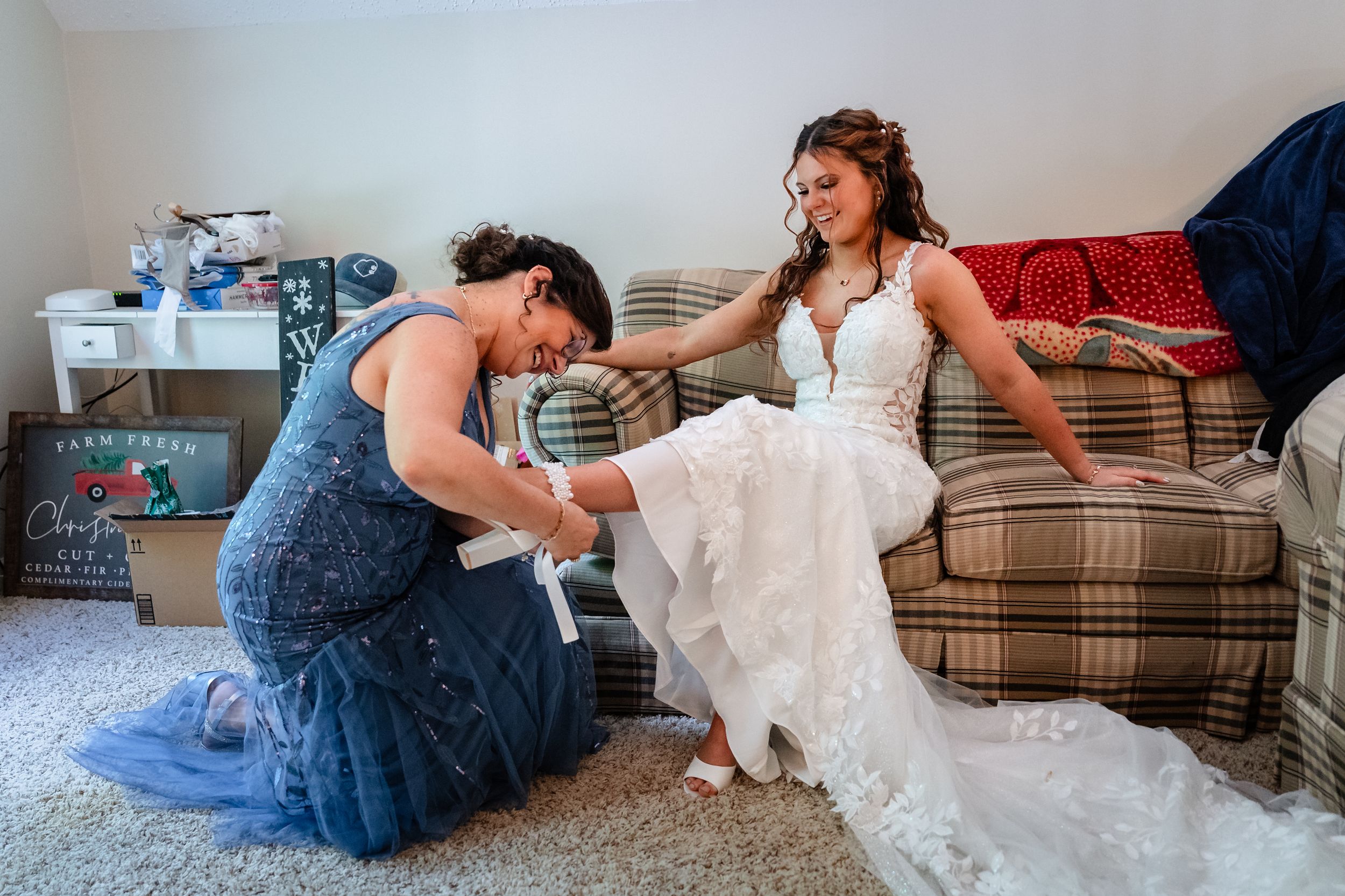 A bridesmaid kneels to help a smiling bride with her shoes while seated on a couch in a wedding dress.