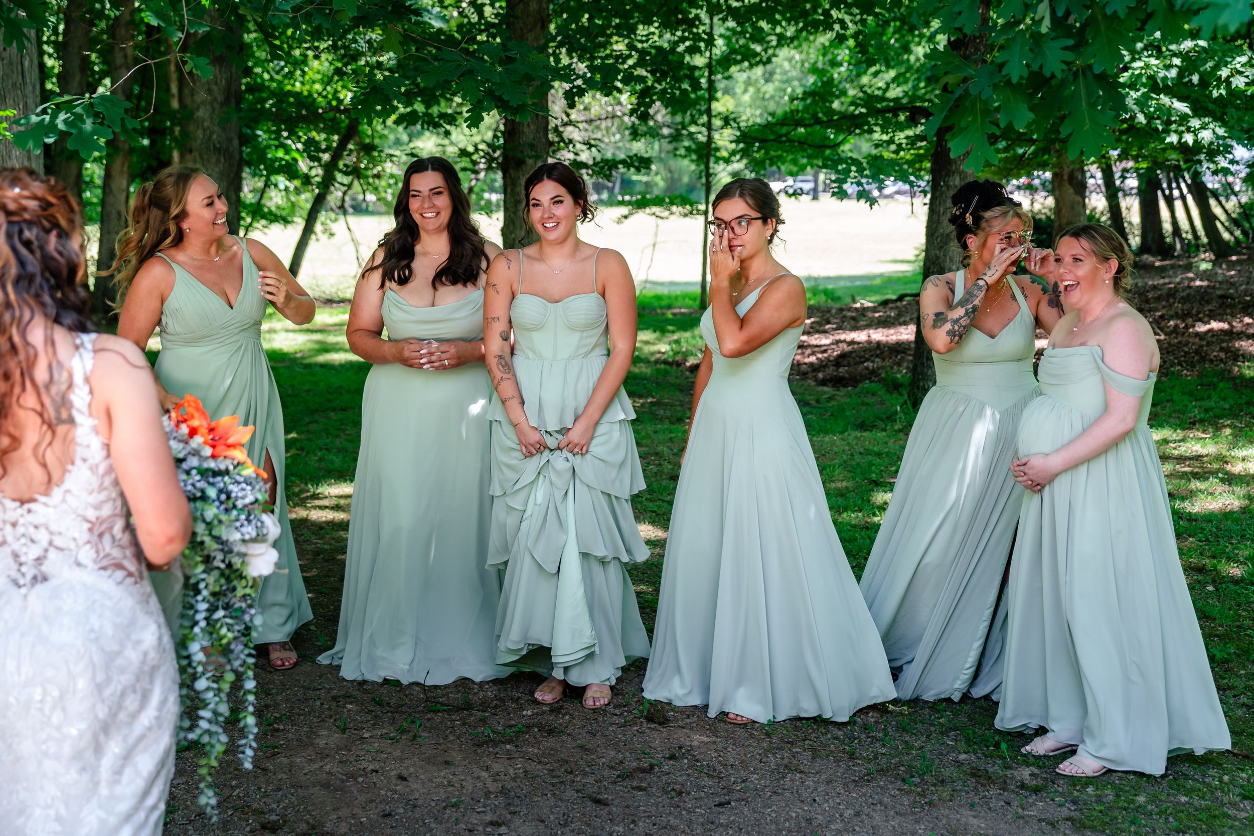 Six bridesmaids in sage green gowns react emotionally as bride approaches for first look outdoors.