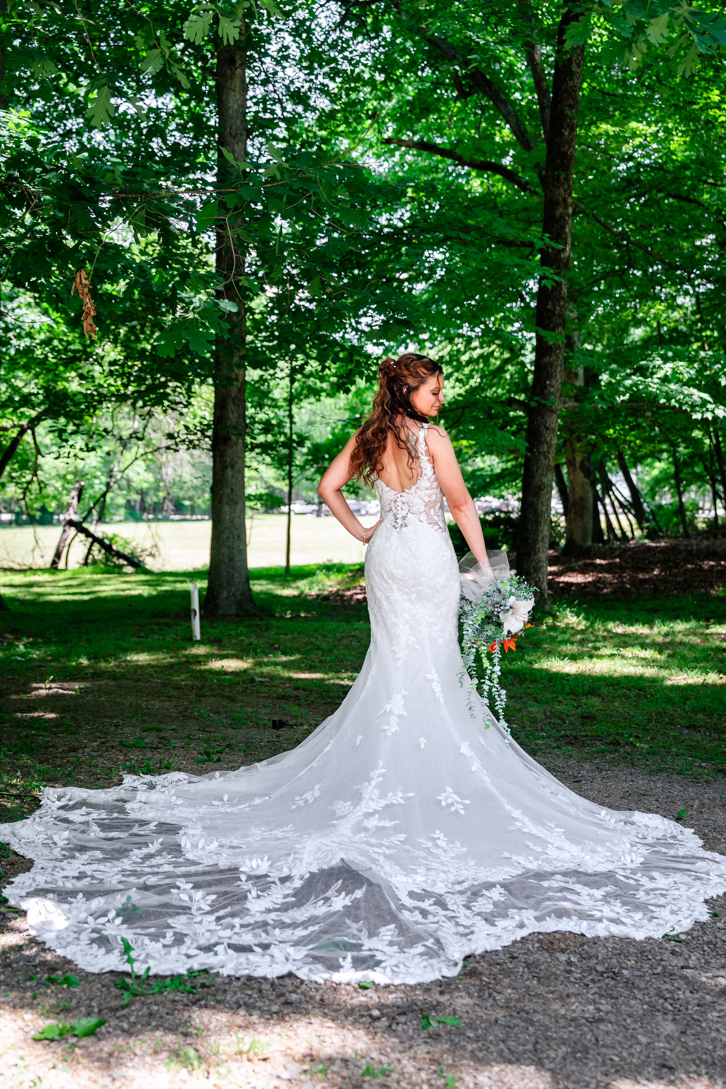 Bride in elegant backless lace mermaid gown with long train posing in green wooded setting.