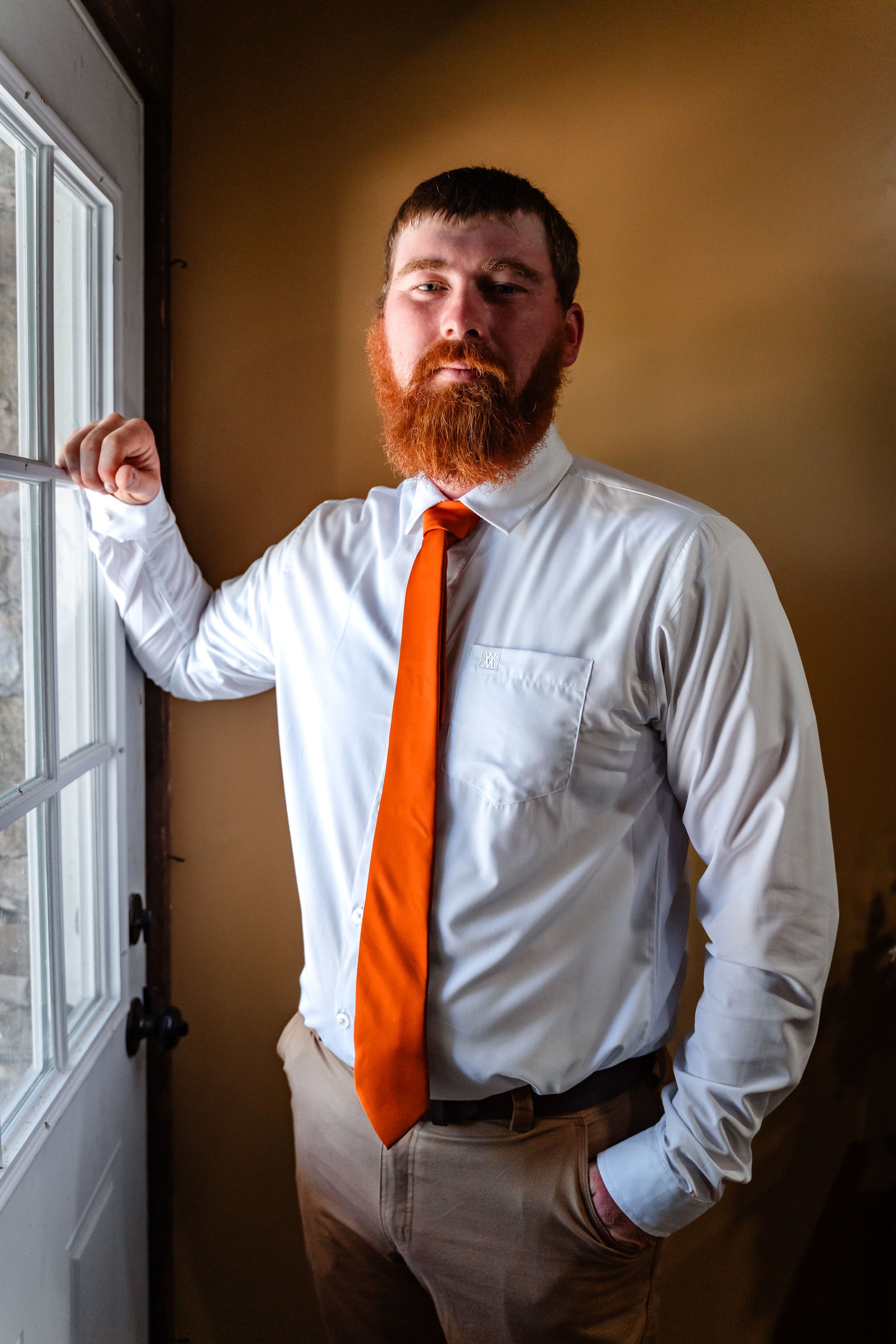 Bearded groom in white shirt and orange tie posing by window in natural light portrait.
