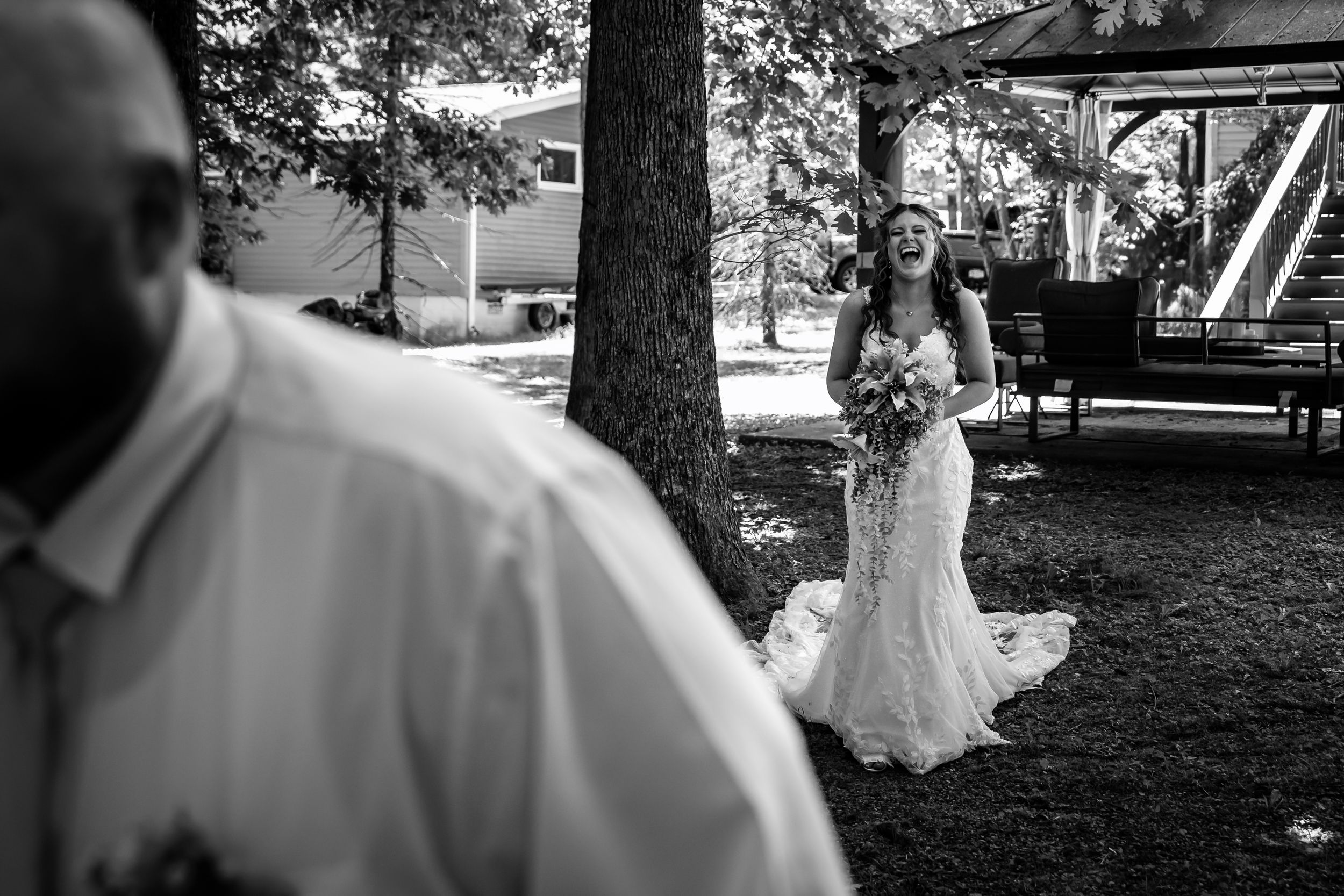 Black and white photo of bride in lace gown approaching groom from behind trees outdoors.