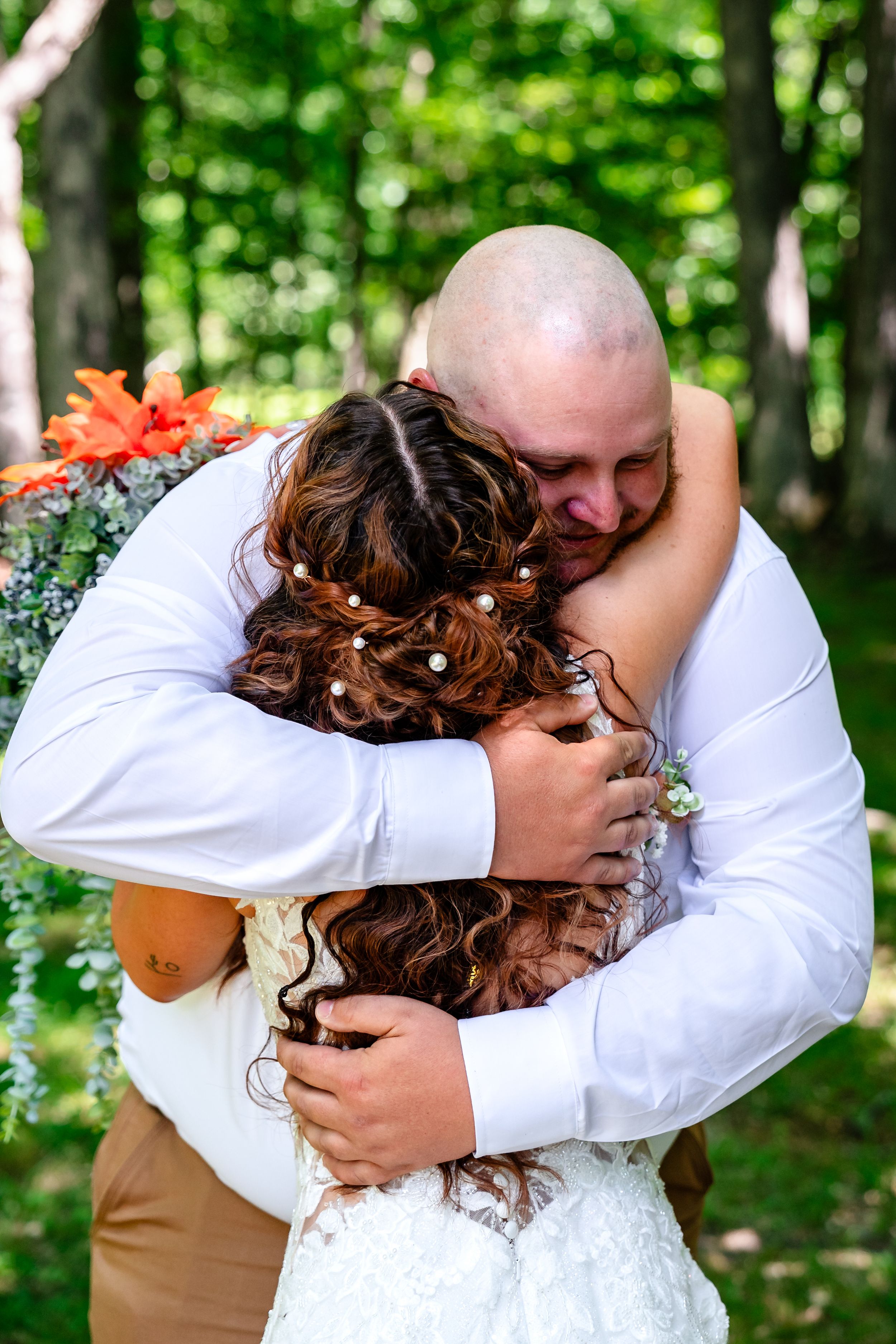 Emotional bald groom embraces his bride tightly during their outdoor wedding ceremony surrounded by greenery.