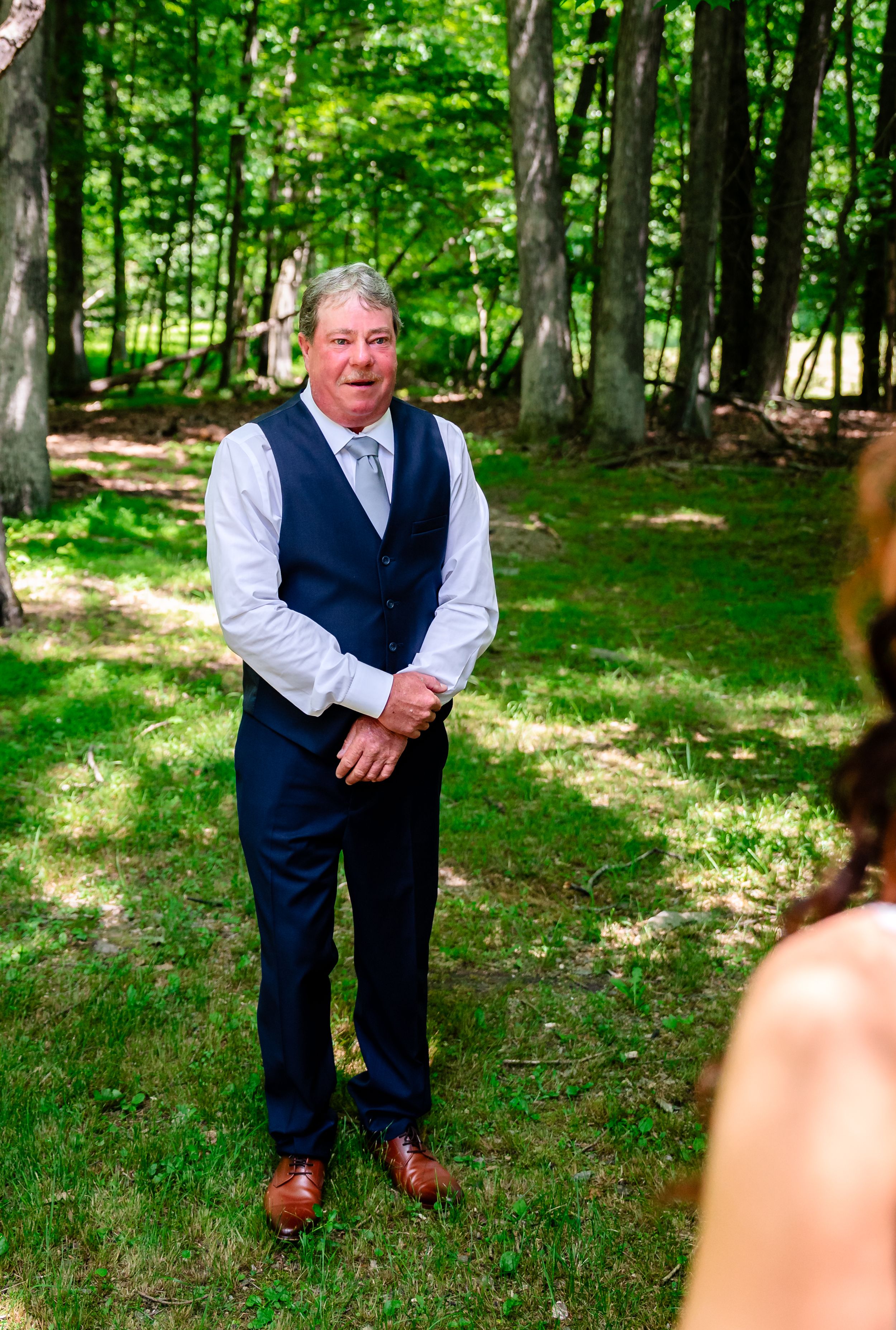 Older man in navy suit vest and tie smiling outdoors at a woodland wedding ceremony.