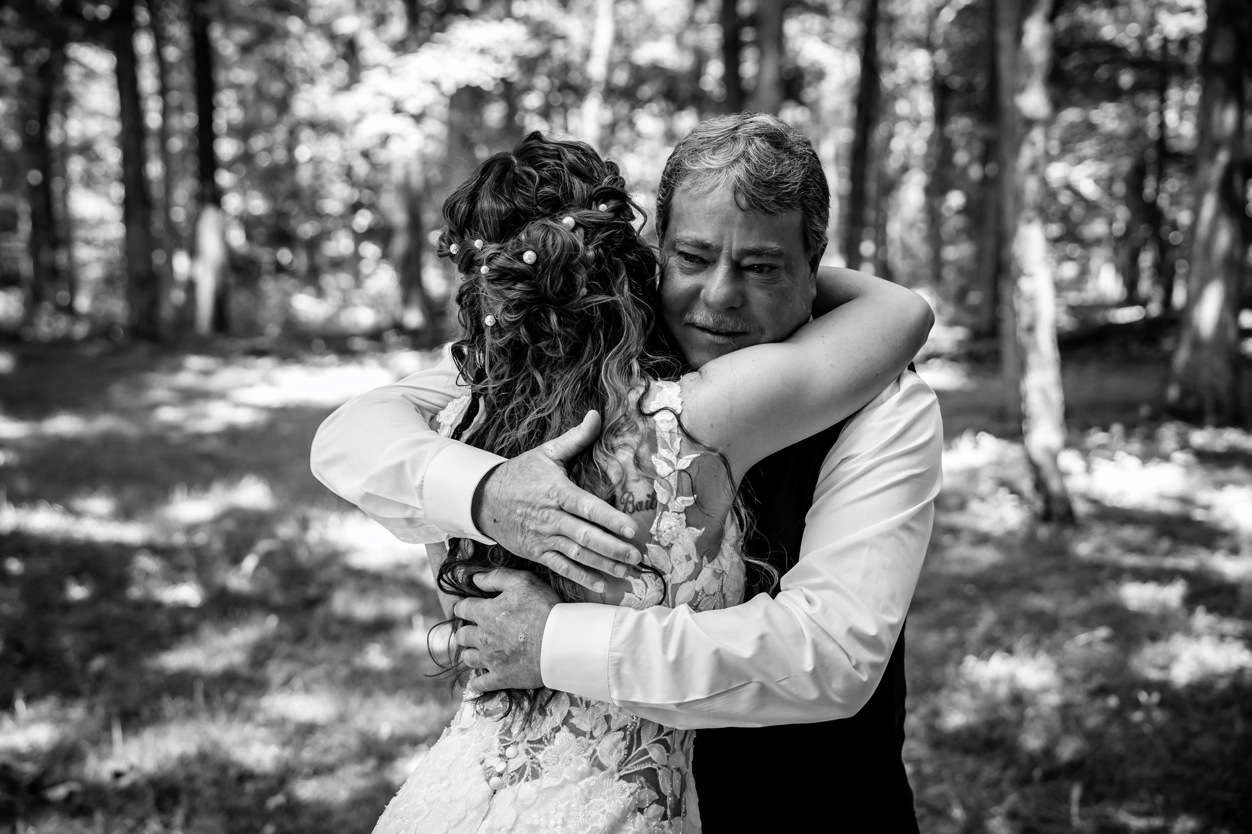 Emotional black and white photo of a bride and older man sharing a heartfelt hug in the woods.