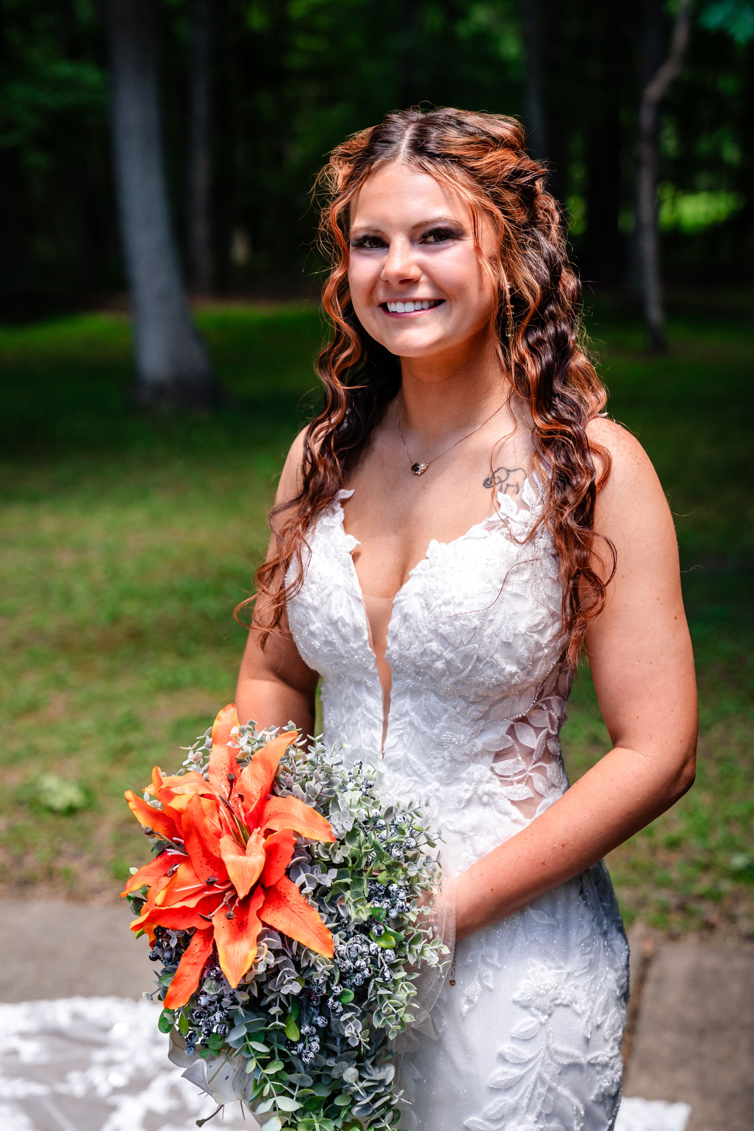 Smiling bride in lace wedding gown holds vibrant orange lily bouquet surrounded by lush green trees.