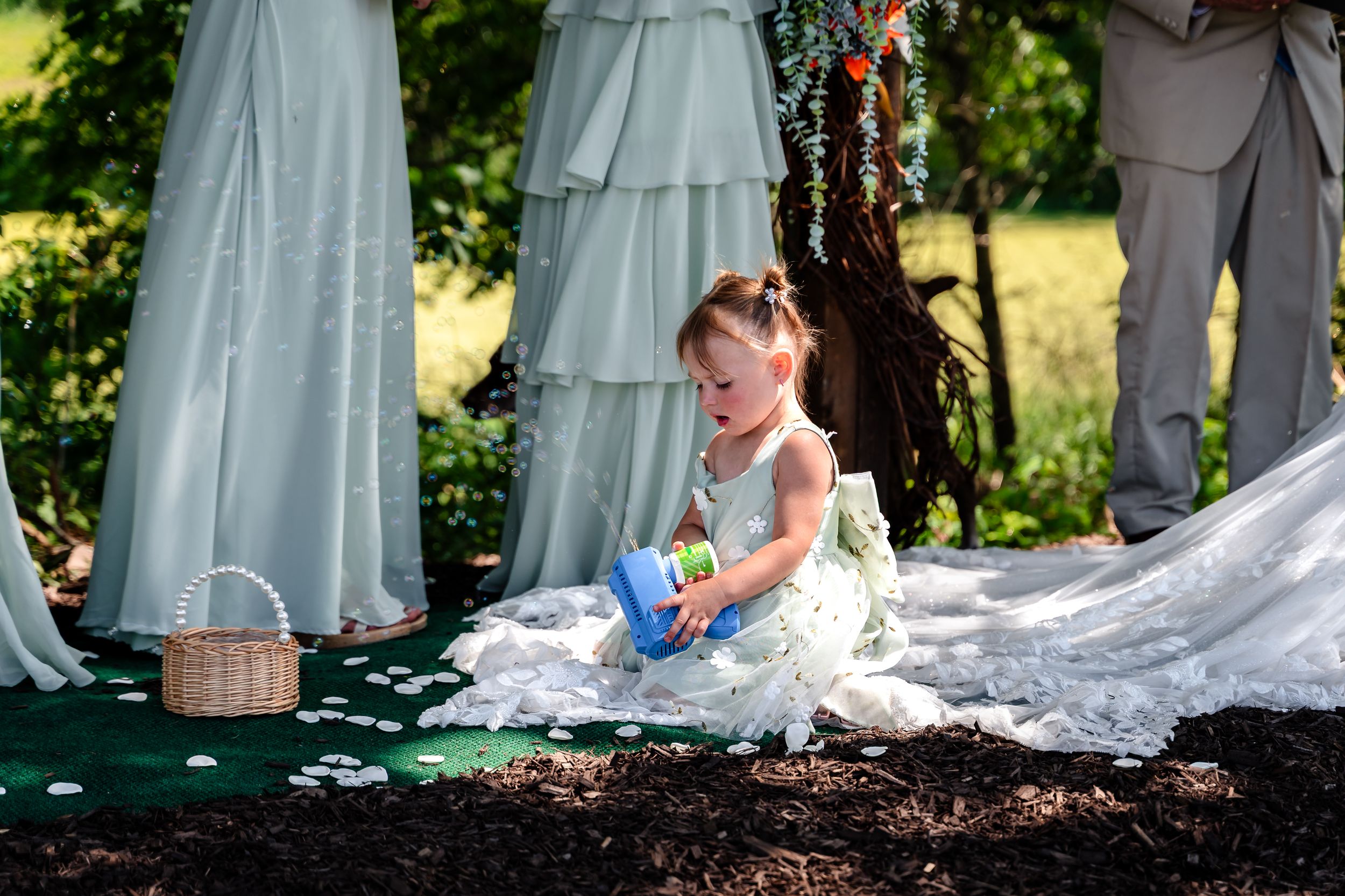Adorable flower girl in white dress playing on wedding train during outdoor garden ceremony.