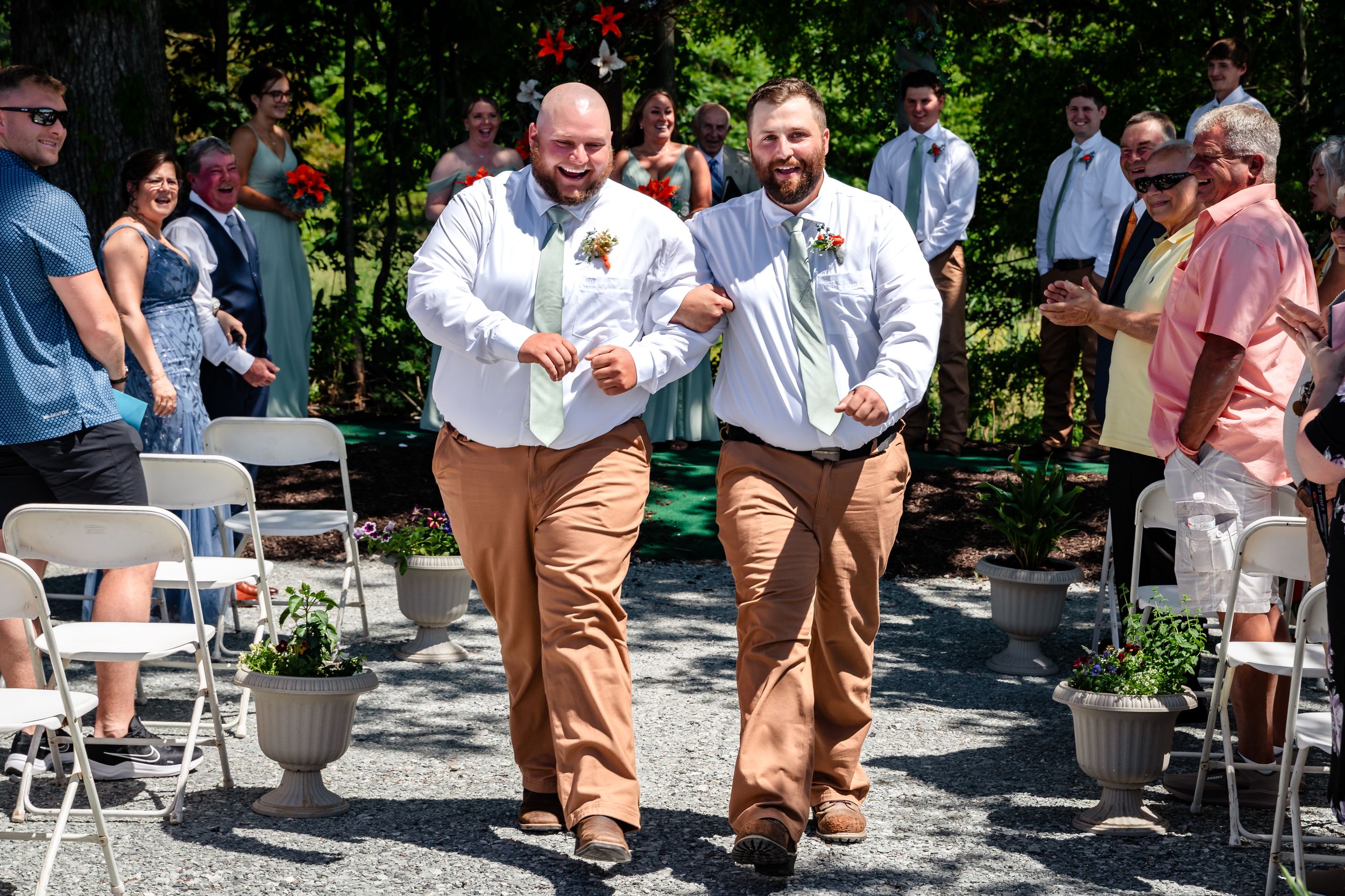 Two smiling groomsmen in white shirts and khaki pants walk down the aisle at an outdoor wedding ceremony.