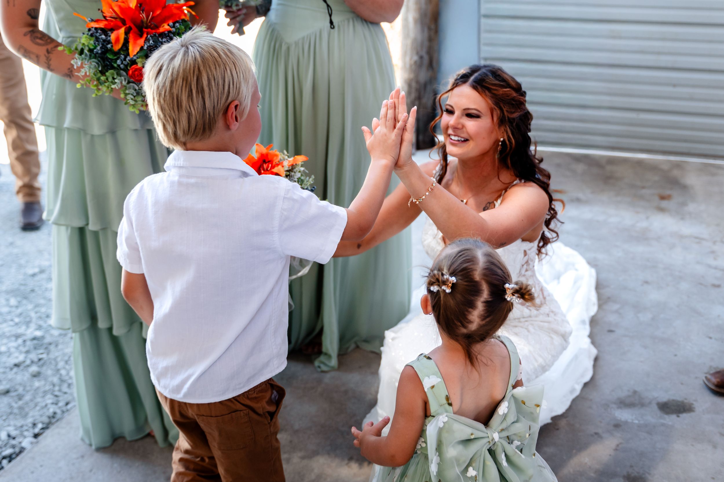 Bride in white gown gives a high-five to a young boy while flower girl watches at wedding.