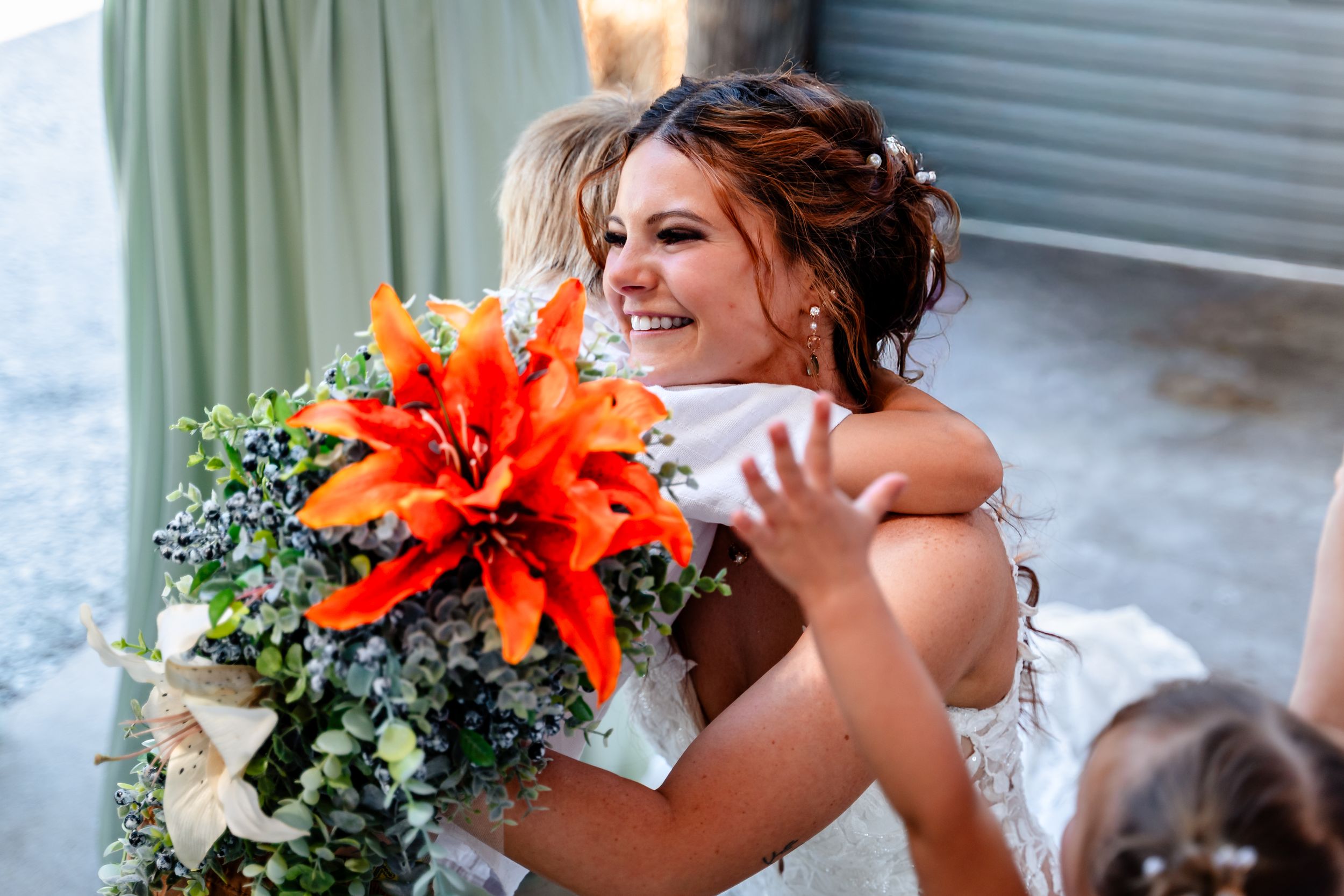 Smiling bride in white dress hugs guest while holding vibrant orange lily bouquet at wedding.