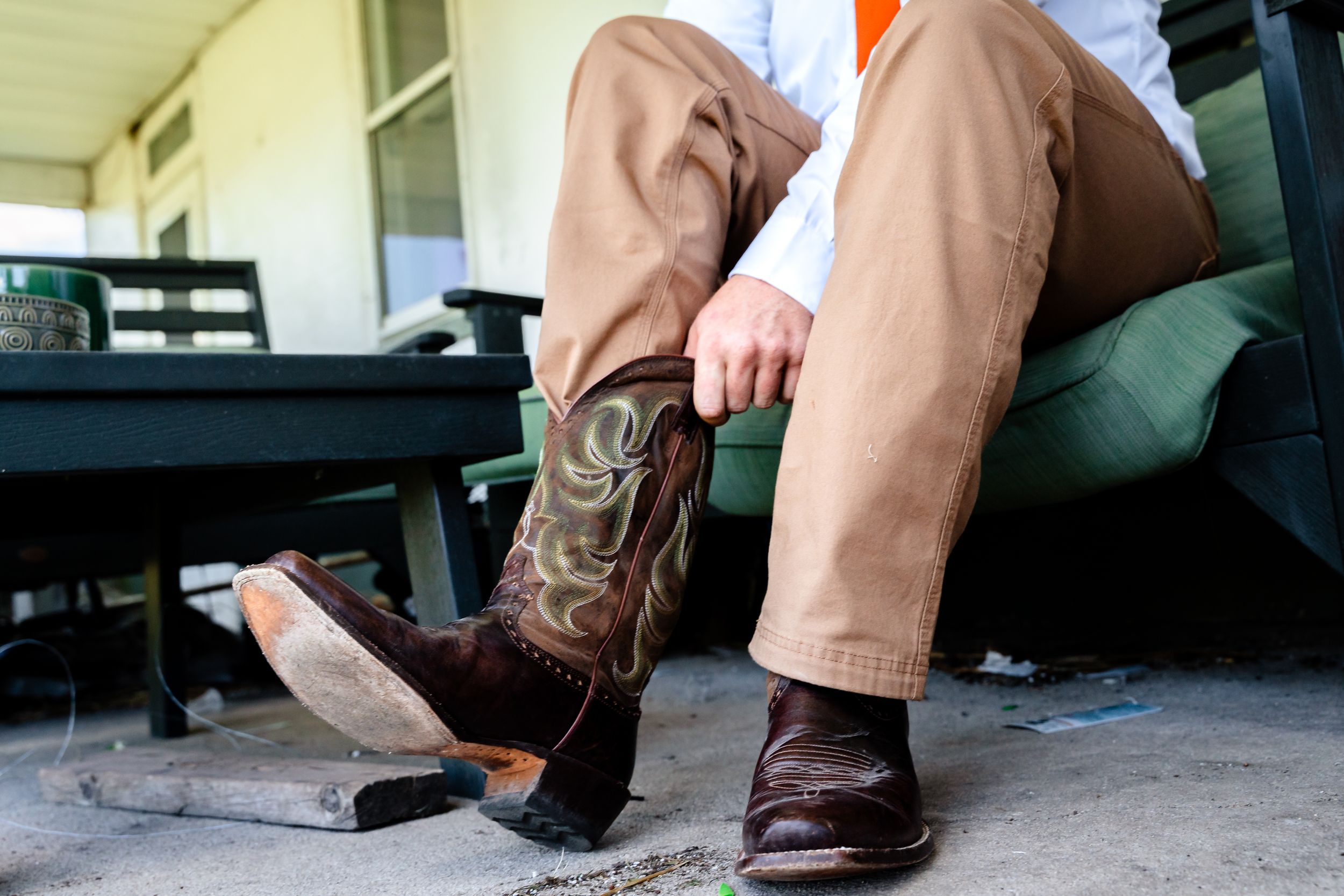 Groom wearing khaki pants puts on brown leather cowboy boots while sitting on a porch before wedding.