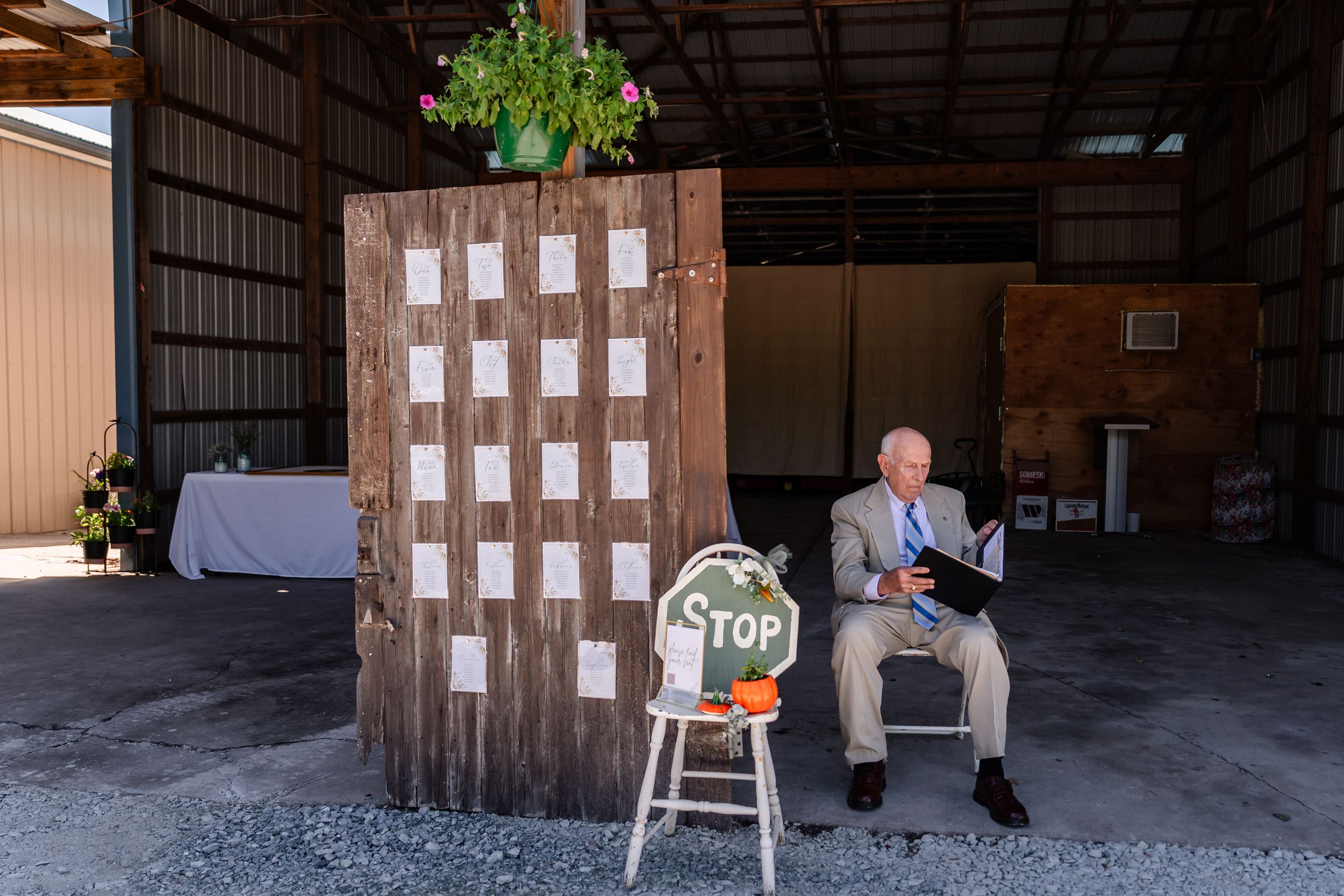 Elderly officiant sits beside a rustic wooden seating chart display inside a barn wedding venue.