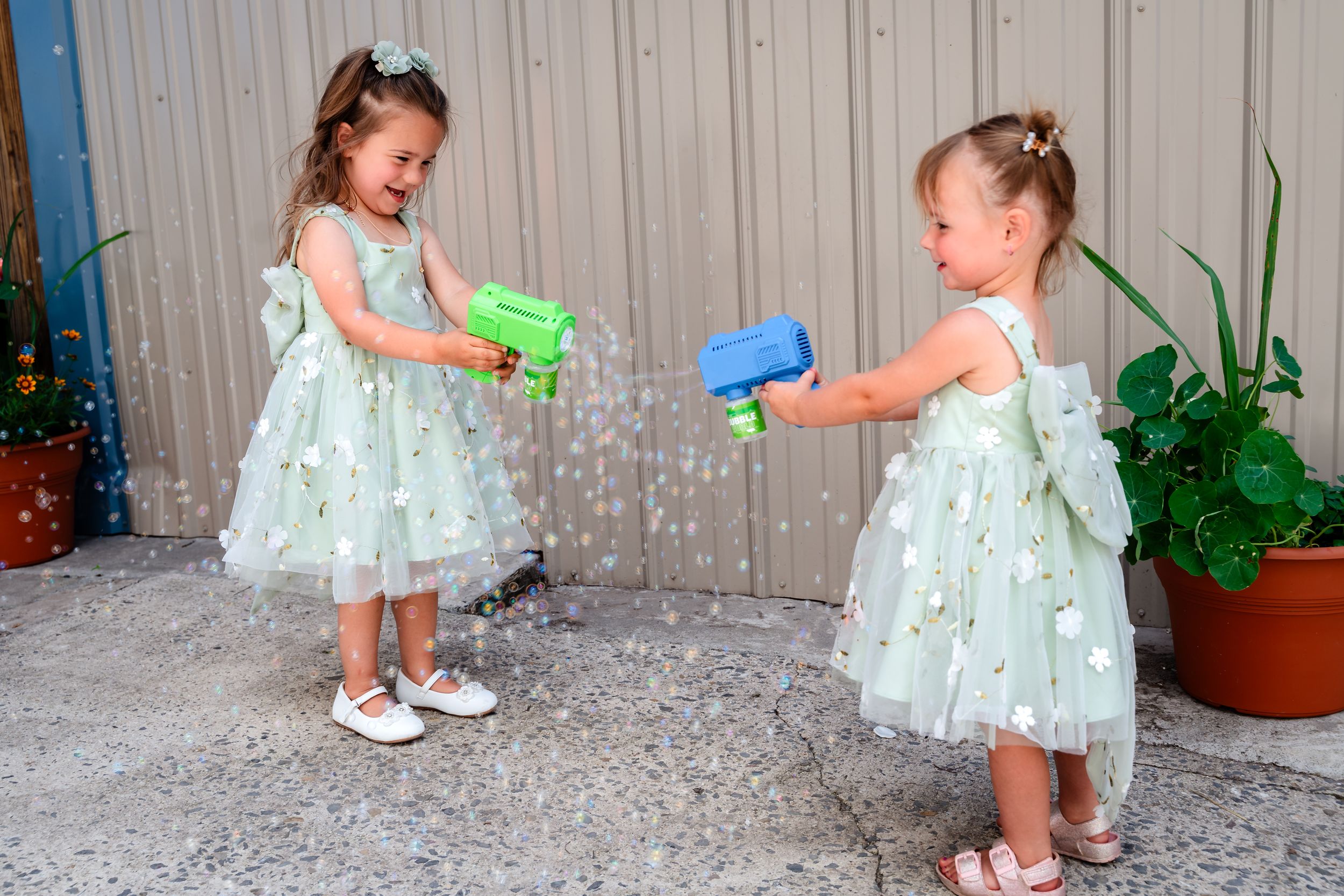 Two little girls in matching green floral dresses play with bubble guns outdoors.