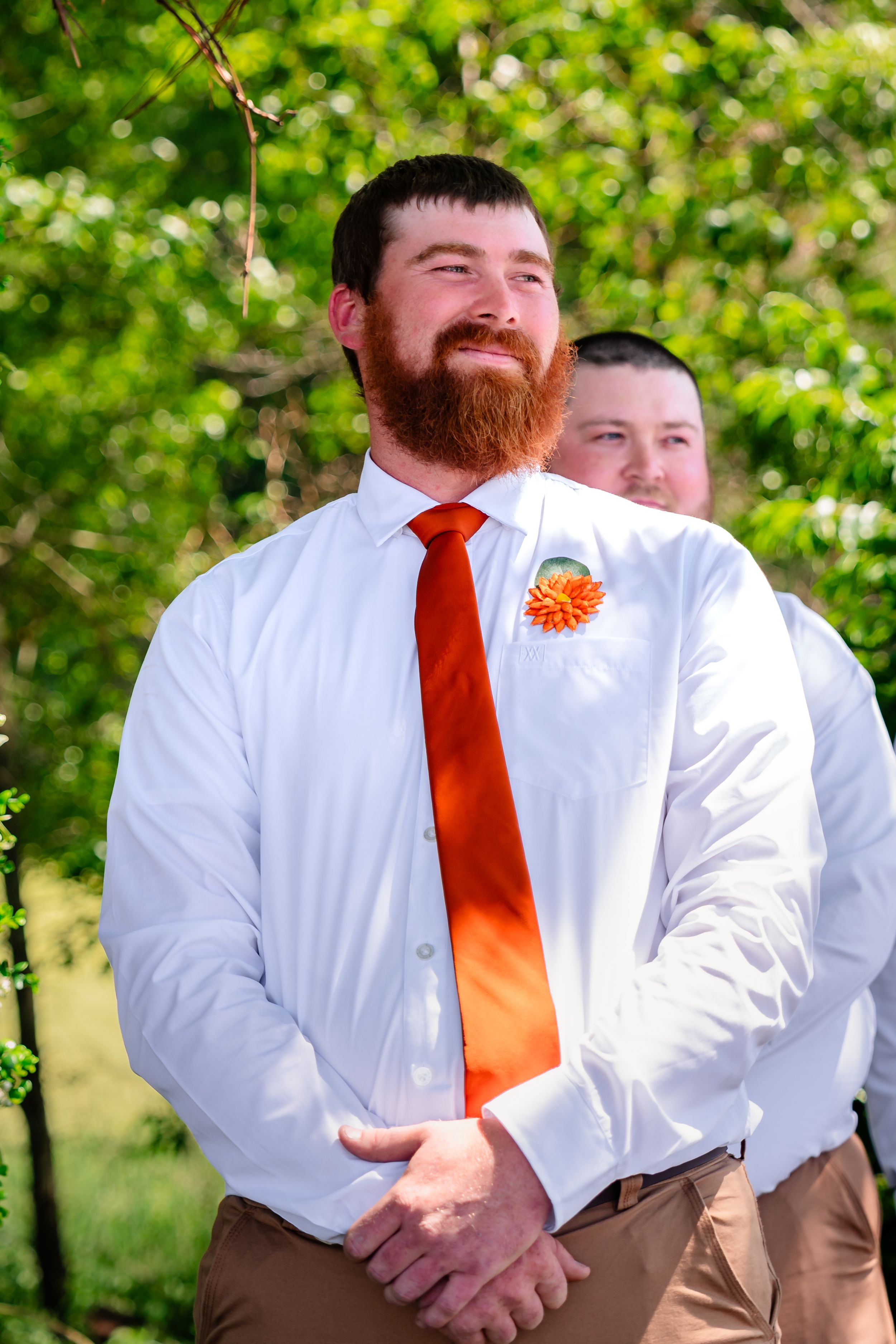 Bearded groom in white shirt and orange tie with boutonniere smiling at outdoor wedding.