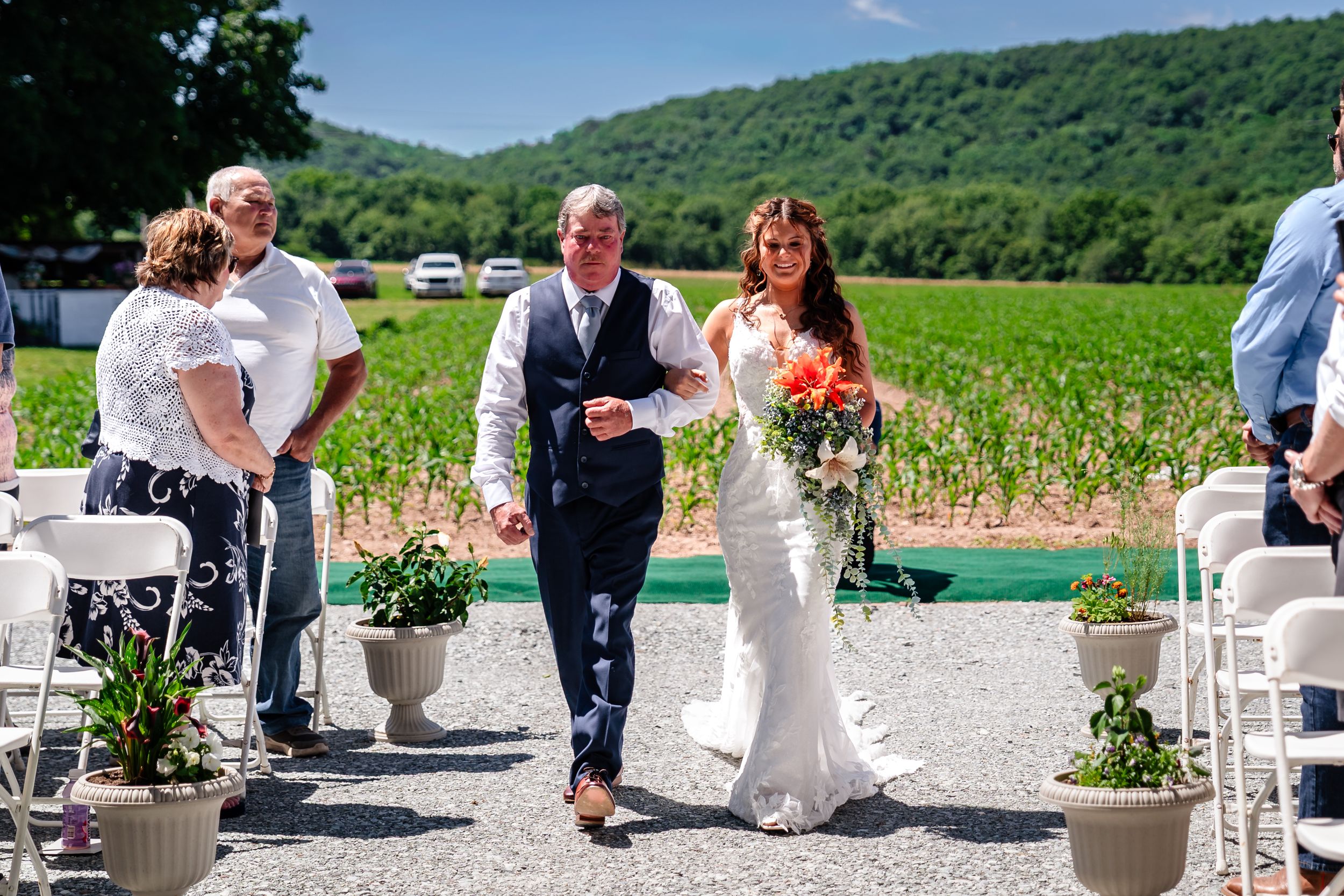 Bride and groom walk down outdoor wedding aisle at vineyard venue on sunny day, bride holding colorful bouquet.