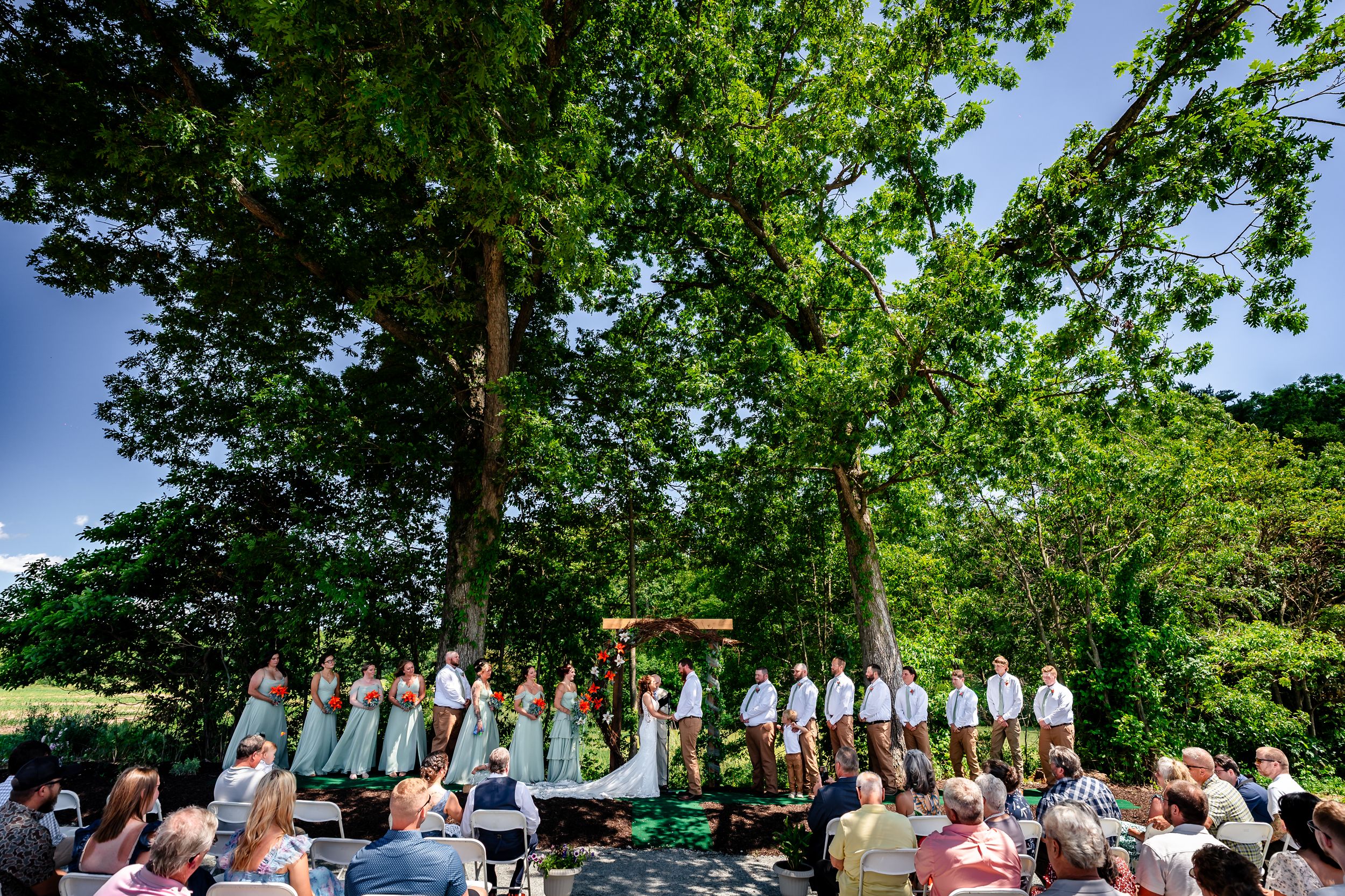 Outdoor wedding ceremony under large oak tree with wedding party in mint green dresses and white suits, guests seated in garden.