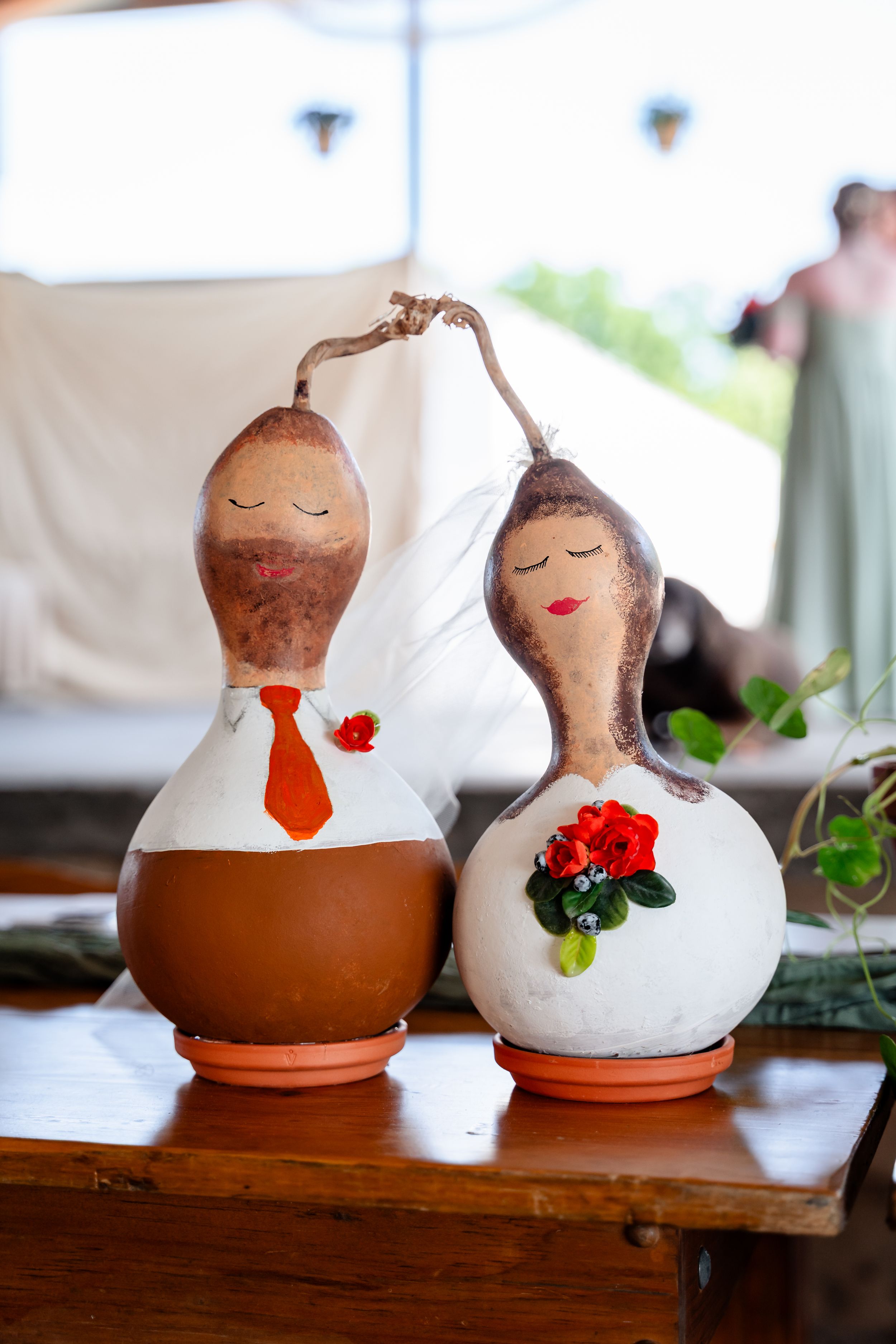 Handmade gourd bride and groom wedding cake toppers with orange tie and red floral details on table.