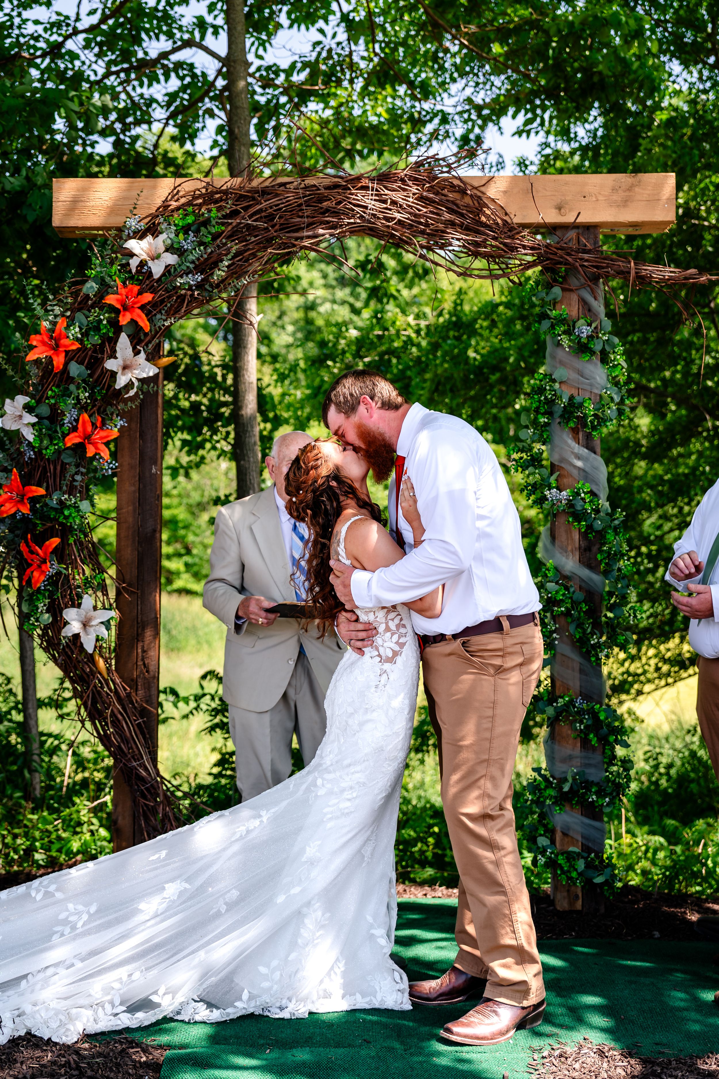 Bride and groom share first kiss under rustic floral arch with orange lilies at outdoor ceremony.