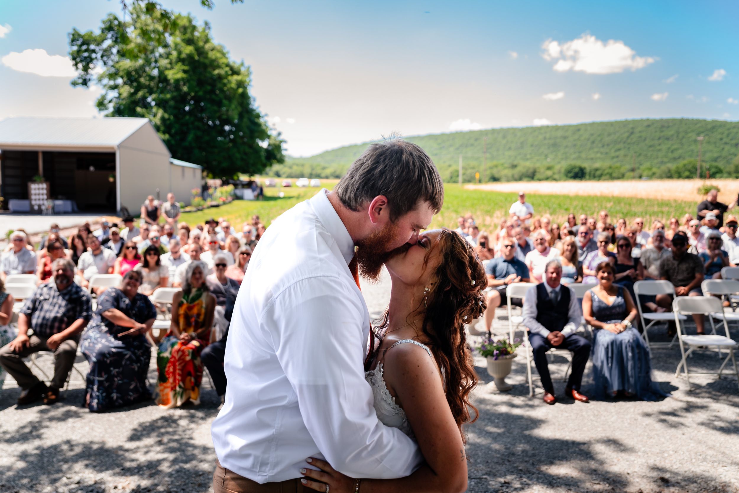 A couple shares their first kiss as newlyweds before a large crowd of guests at an outdoor wedding ceremony.
