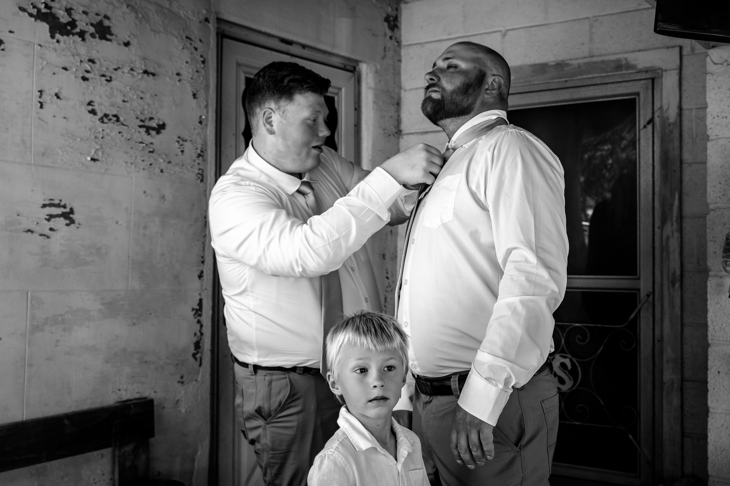 Black and white photo of groomsmen helping groom with tie as young boy stands between them.