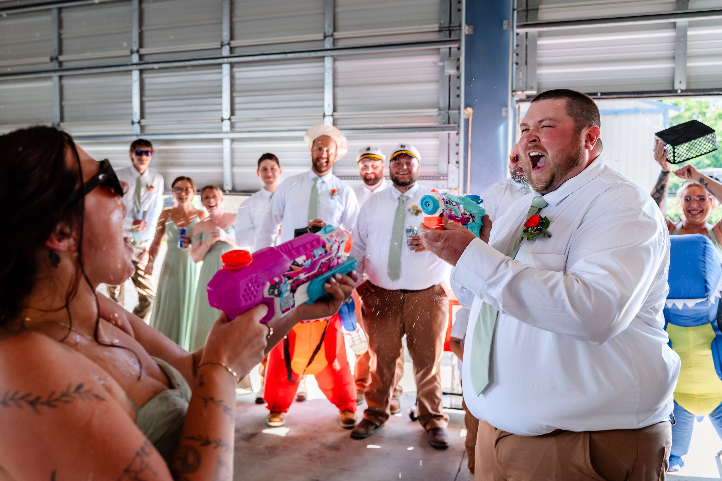 Laughing groom holds colorful water guns as bridal party surprises him inside a barn at the wedding reception.