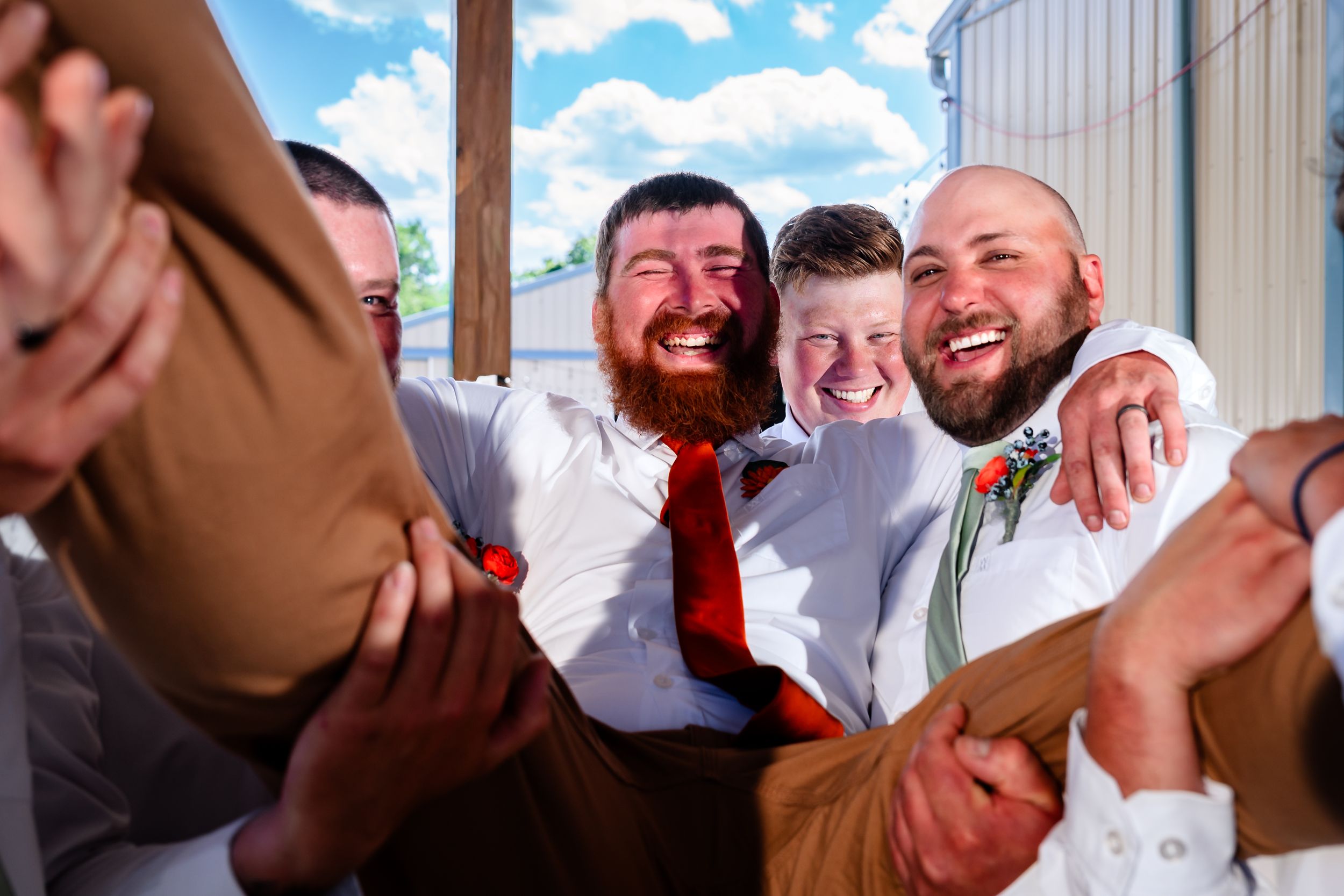 Joyful groomsmen laugh and celebrate together outside on a sunny wedding day, one wearing a red tie.