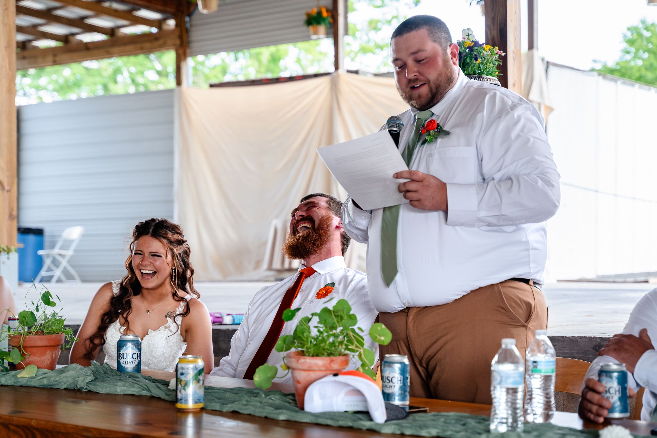 Best man delivers humorous wedding speech as bride and groom laugh at head table during reception.