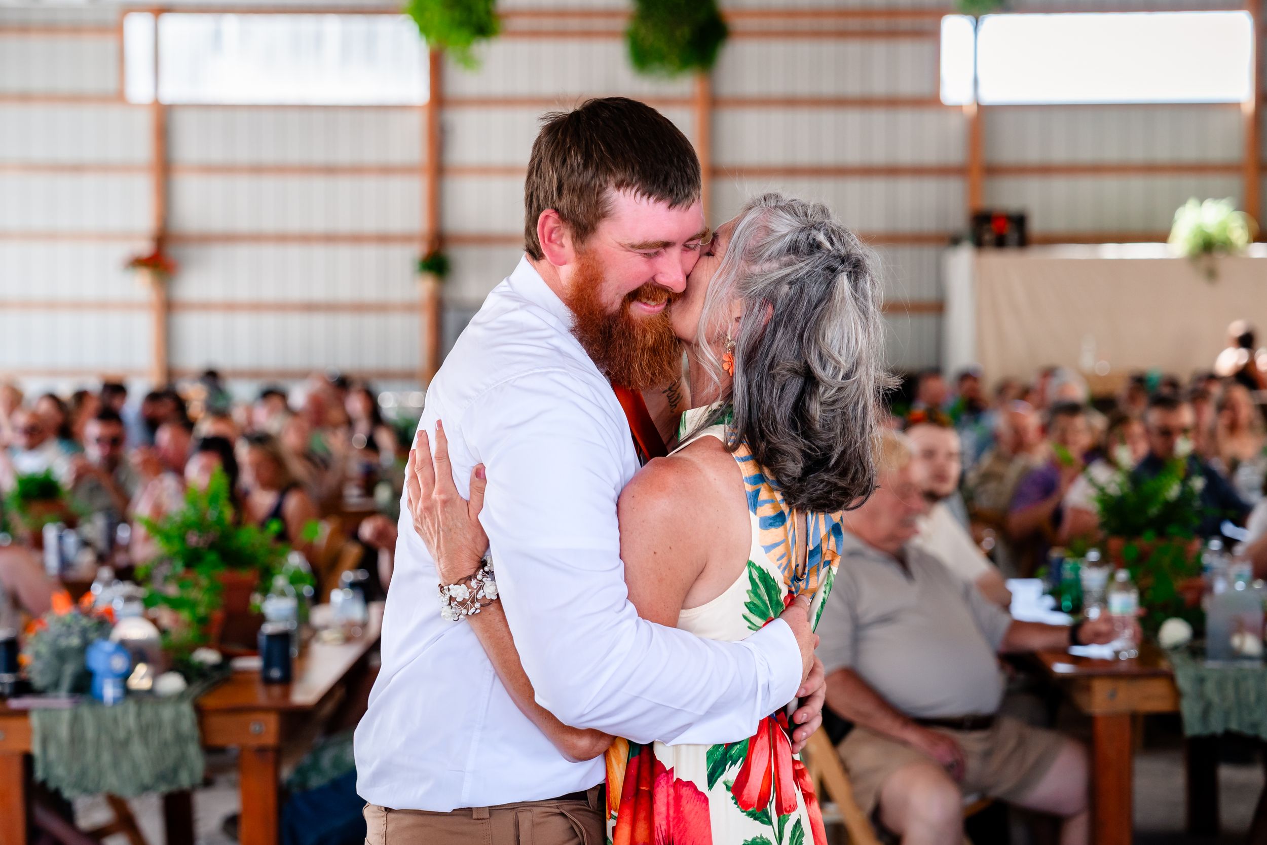 Groom with red beard shares a warm mother-son dance at a crowded barn wedding reception.