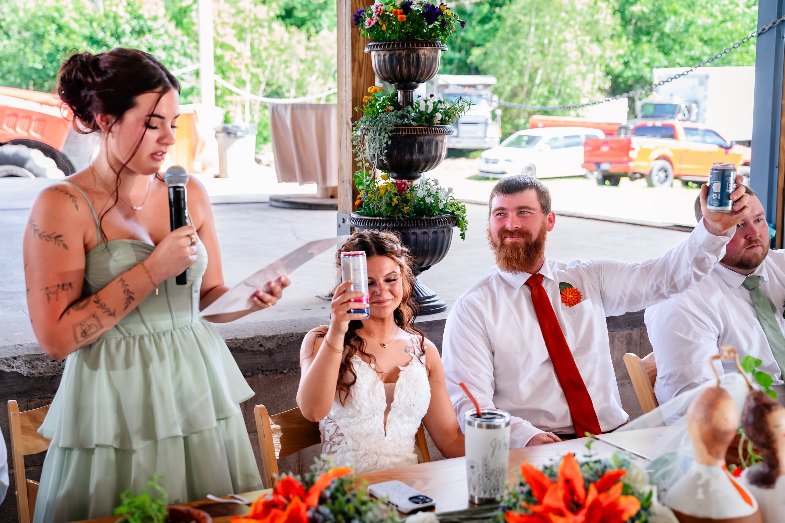Bride and groom celebrate at wedding reception as bridesmaid gives a toast, holding drinks and laughing at decorated table.
